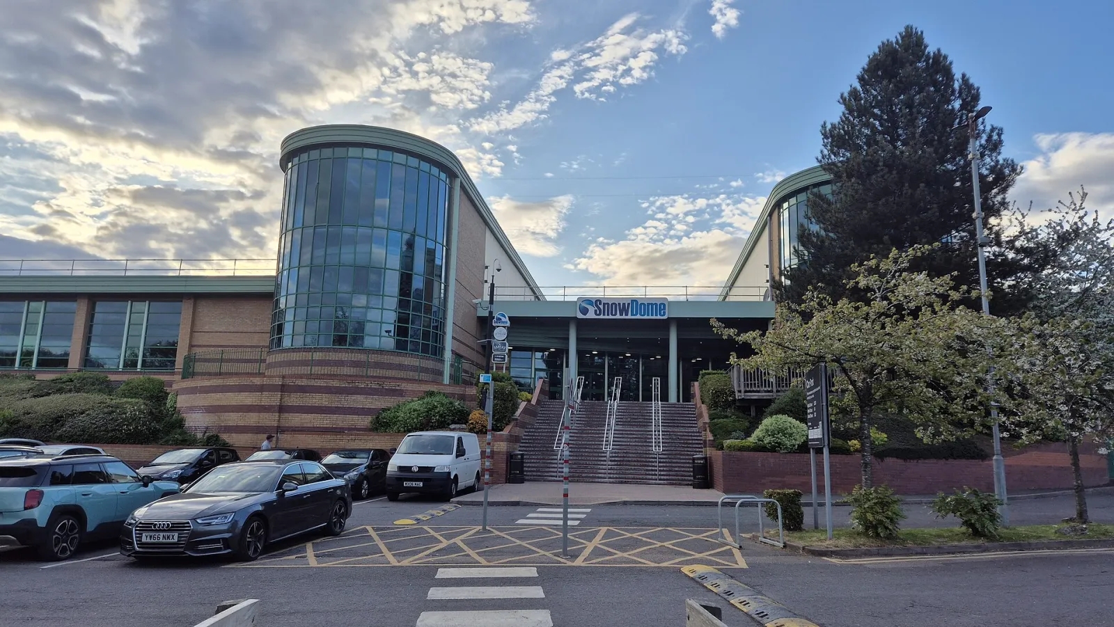 The front of the SnowDome building in Tamworth: a glazed round tower and the covered main entrance with its steps, seen across the car park. — SnowDome (Tamworth)