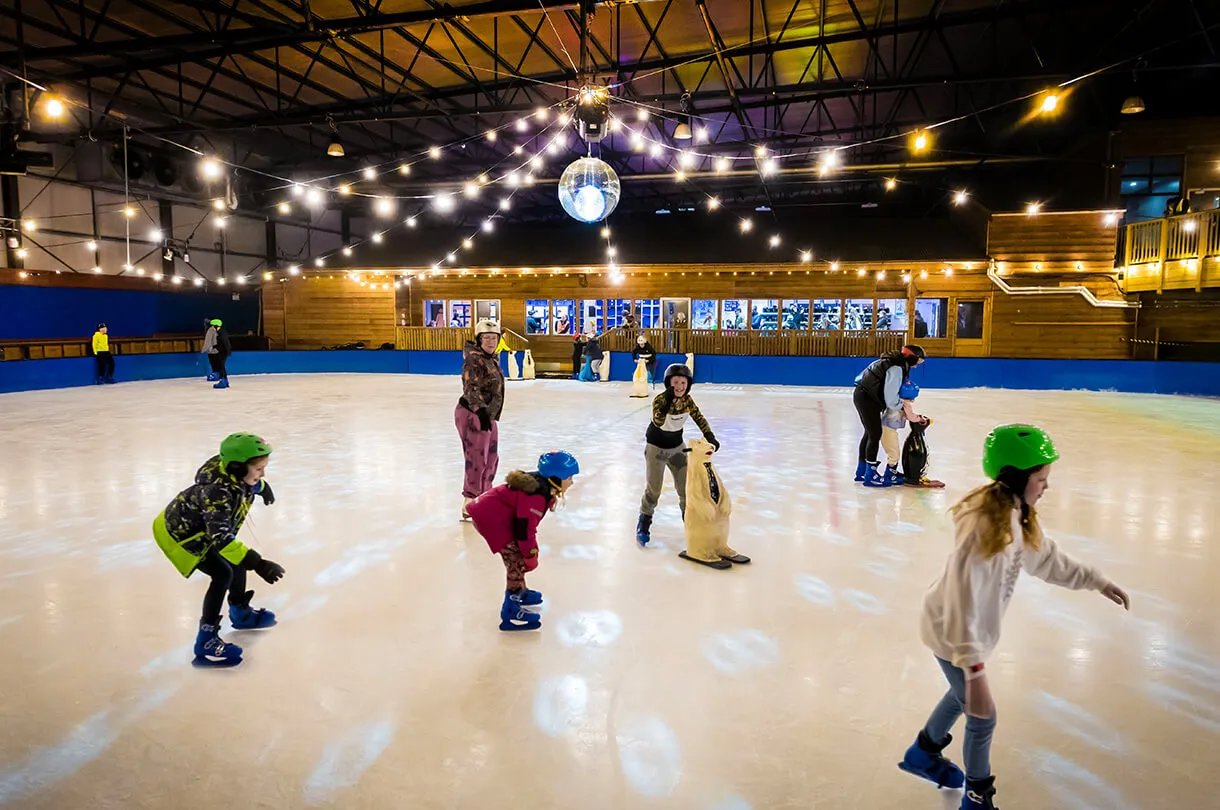 The SnowDome ice rink with skaters spread across the ice under strings of lights, wooden lodge and boards behind, several children on the ice with skating aids. — SnowDome (Tamworth)