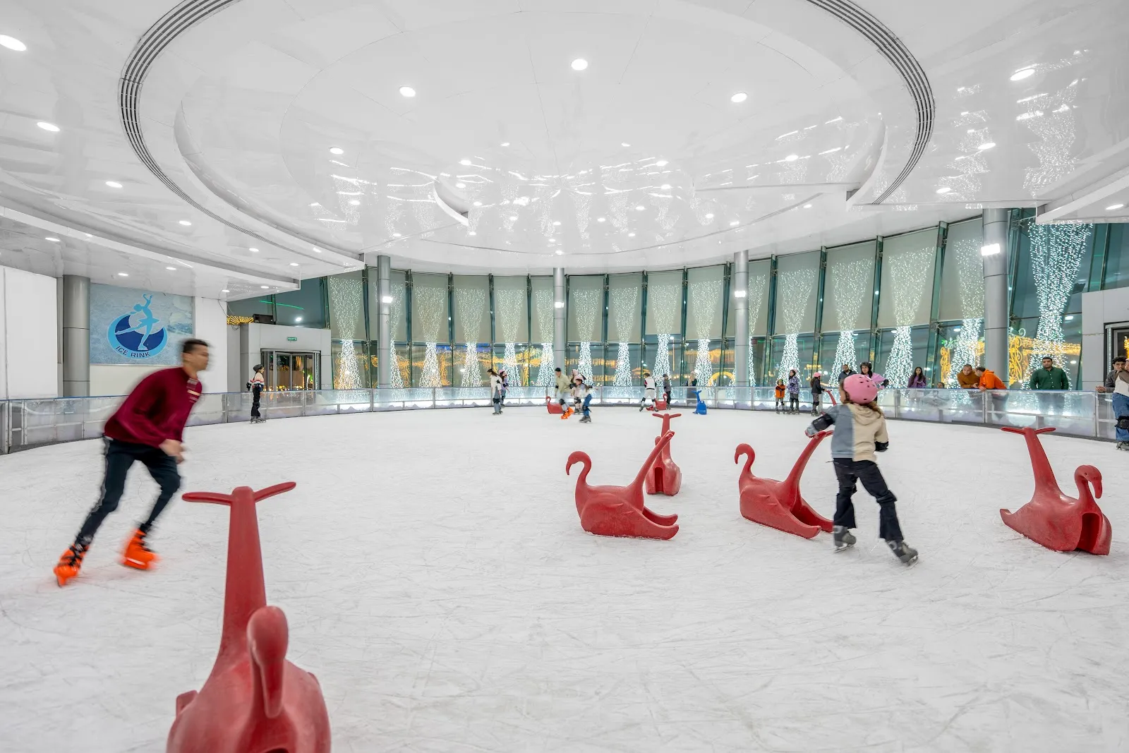 Skaters and red seal-shaped skate aids on the ice of the SOHO Square rink, in a round hall with a lit ceiling and a glass wall of fairy lights. — Ice Rink at SOHO Square