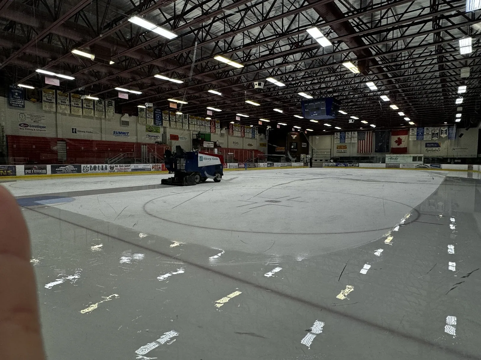 La pista de tamaño olímpico con la máquina alisadora trabajando y los asientos y banderas con ribete rojo en la pared del fondo. — Soldotna Ice Arena