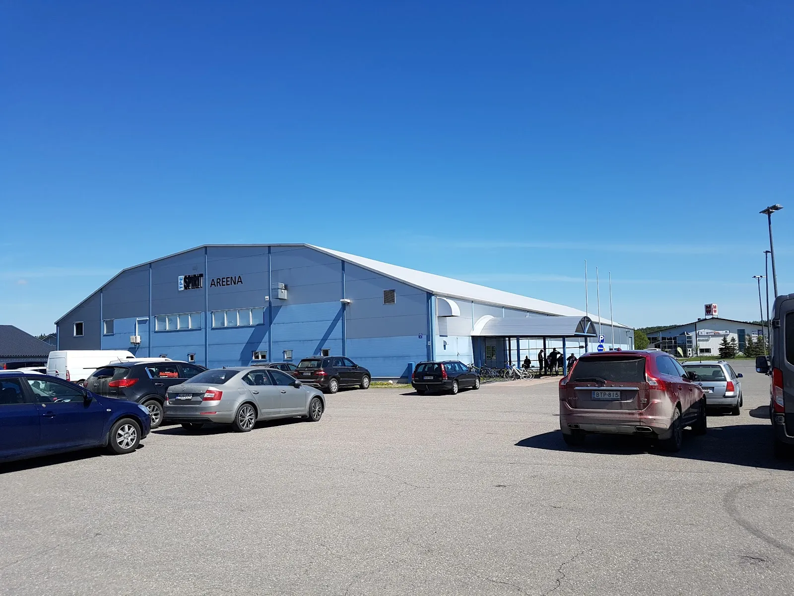The pale blue-roofed Somero ice hall behind a gravel car park with several cars parked in front. — Someron jäähalli