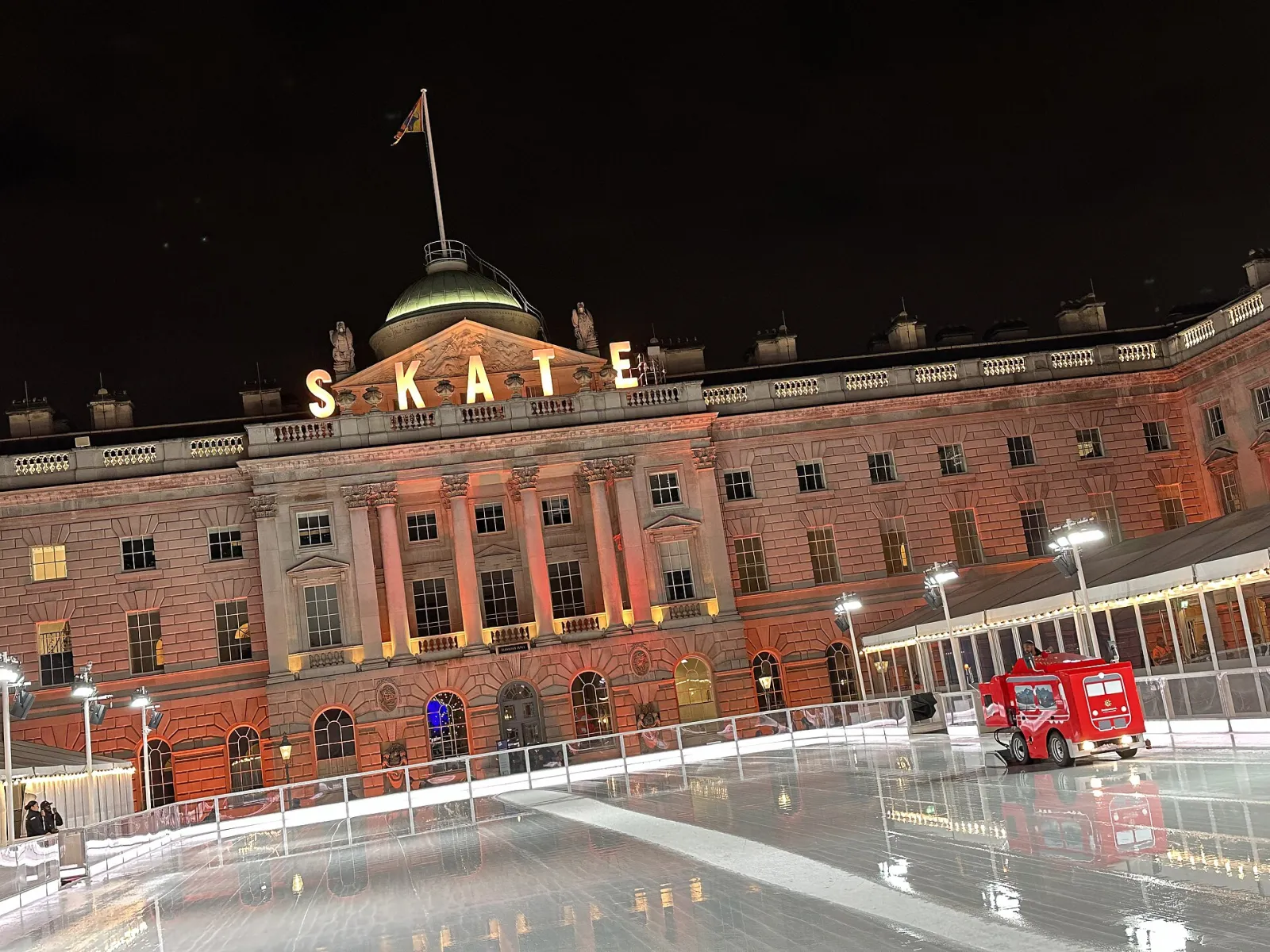 Somerset House's domed courtyard front at night, lit orange with SKATE letters on the roofline, above the empty rink and an ice resurfacer. — Skate at Somerset House