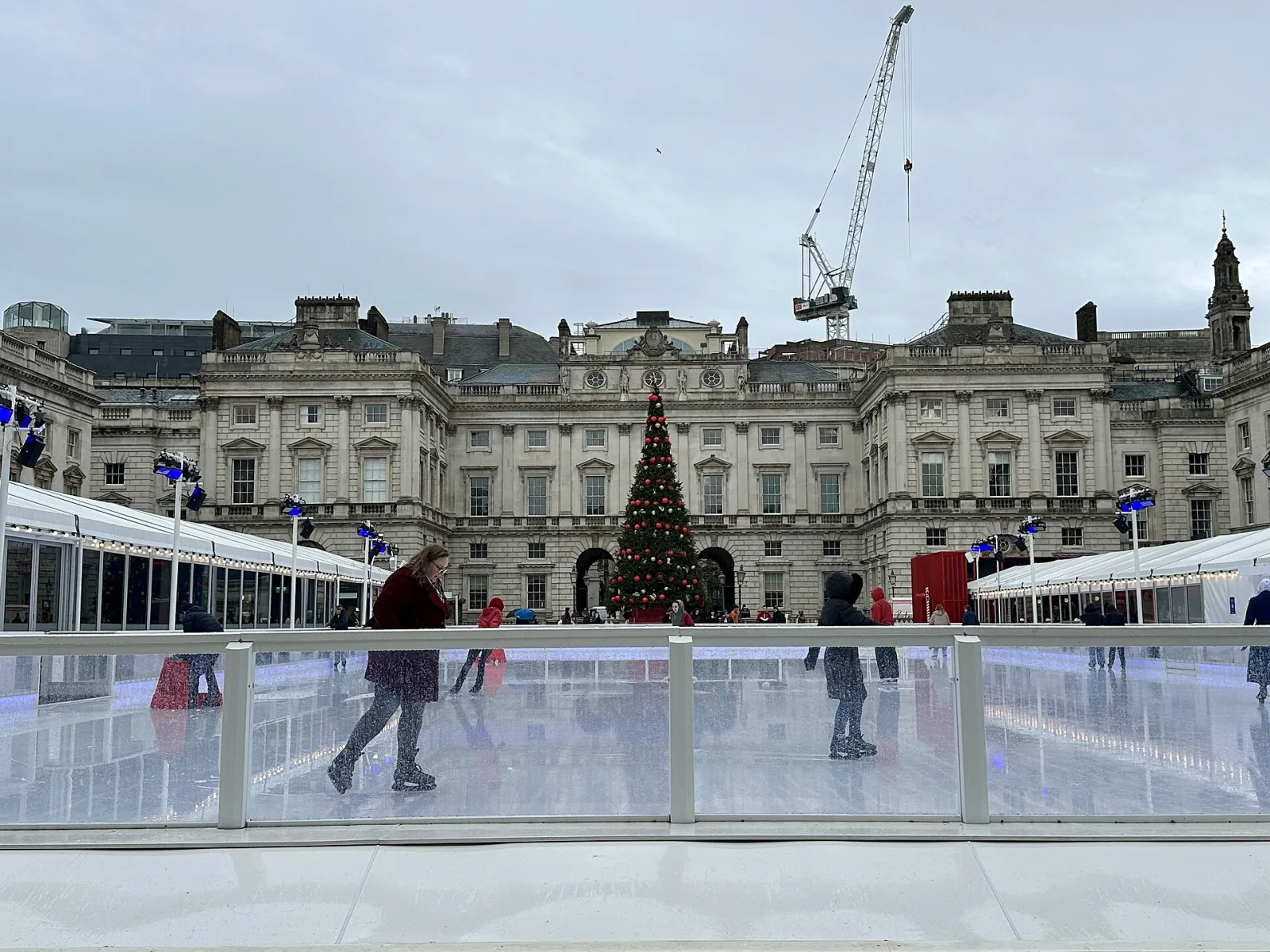 The courtyard rink by day with a few skaters, a Christmas tree and the Somerset House facade behind the rink barrier. — Skate at Somerset House