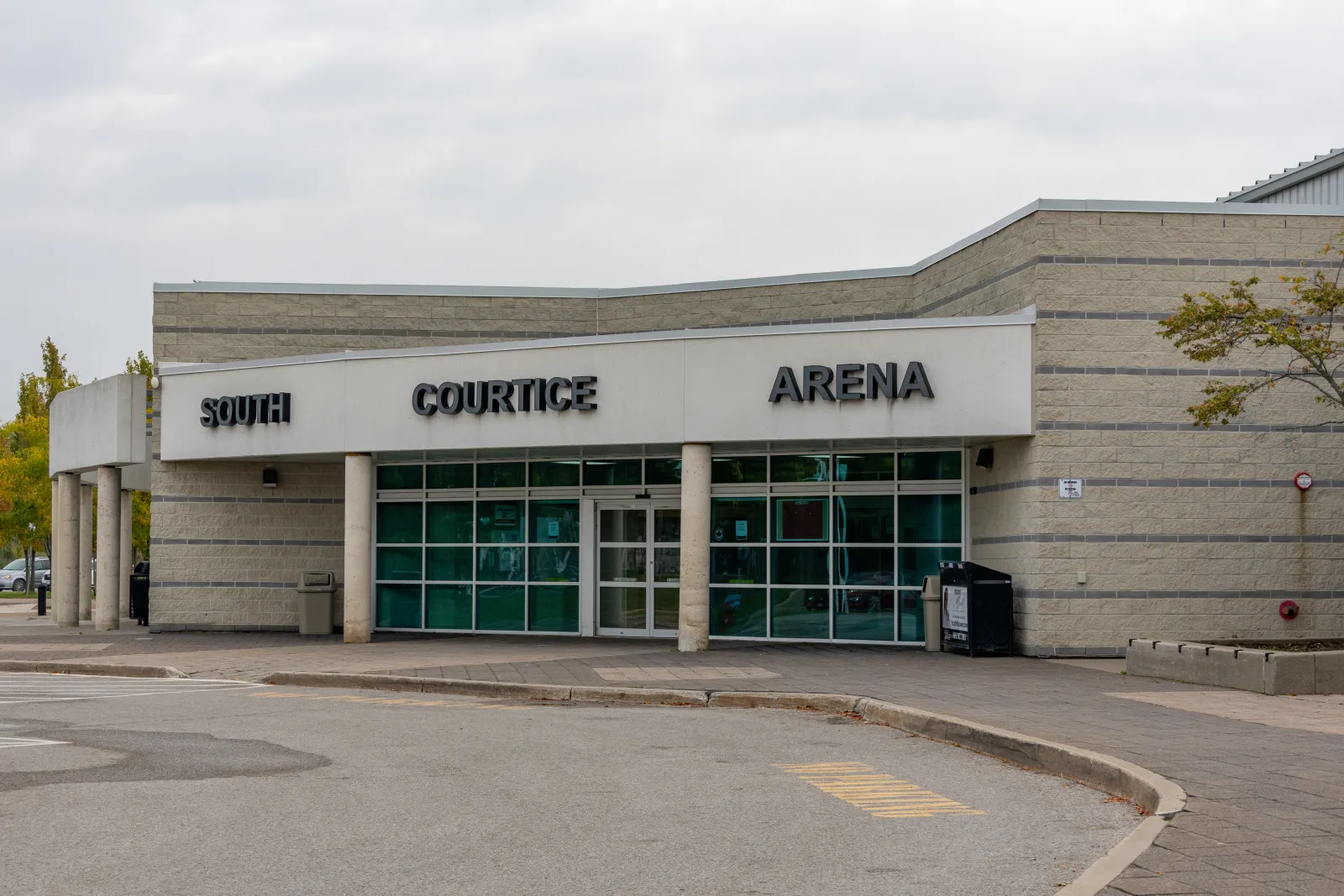 La entrada principal de South Courtice Arena: una fachada de bloques claros con "South Courtice Arena" en letras negras sobre un vestíbulo de cristal, con el carril circular para dejar pasajeros al frente. — South Courtice Arena