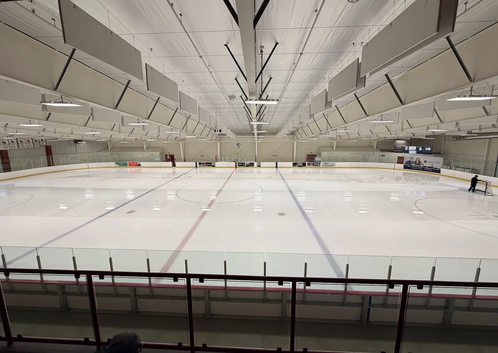El hielo vacío visto desde el barandal superior, con líneas rojas y azules bajo un techo blanco del que cuelgan hileras de paneles acústicos. — South Courtice Arena