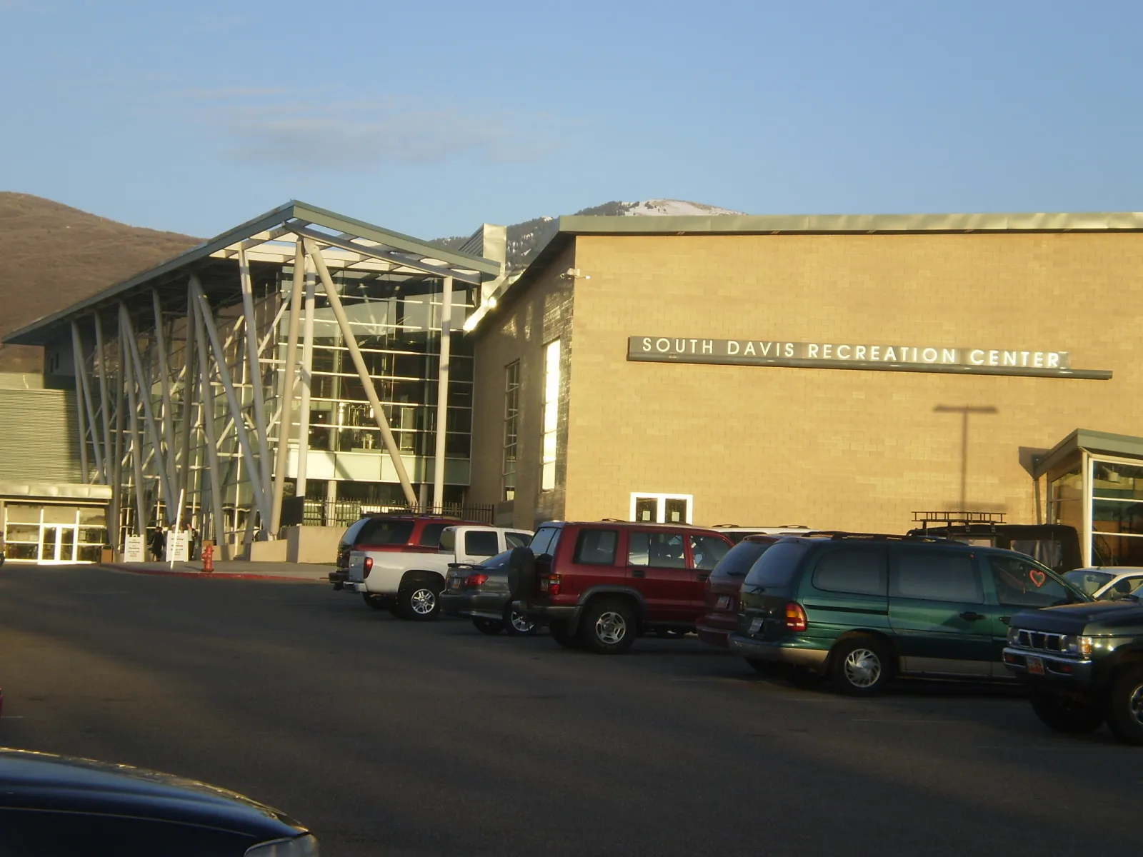 El edificio desde el estacionamiento, con el nombre South Davis Recreation Center en el muro de ladrillo junto a la entrada de vidrio y sus columnas de acero inclinadas. — South Davis Recreation Center