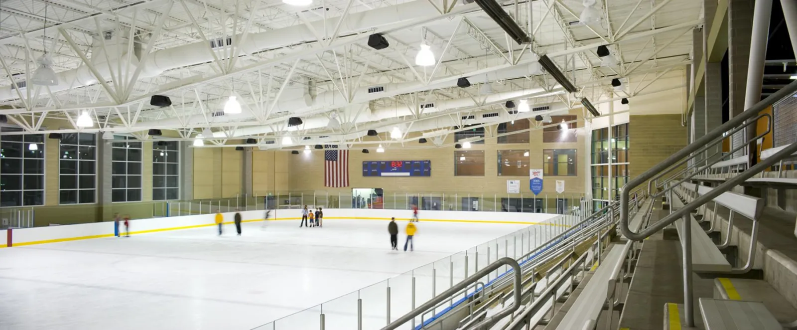 La pista vista desde lo alto de las gradas, con algunos patinadores en el hielo, ventanales altos en la esquina del fondo y una bandera en el muro del extremo. — South Davis Recreation Center