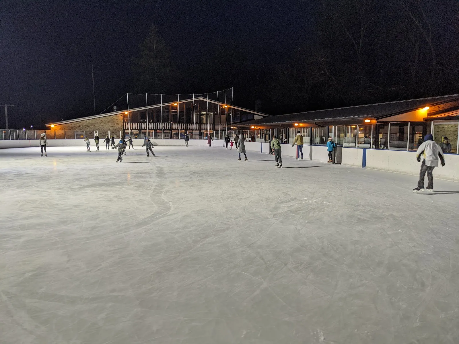 The outdoor rink under the lights on a winter evening, a wide sheet of ice with skaters near the far boards and the low rink lodge glowing along one side. — South Park Ice Rink
