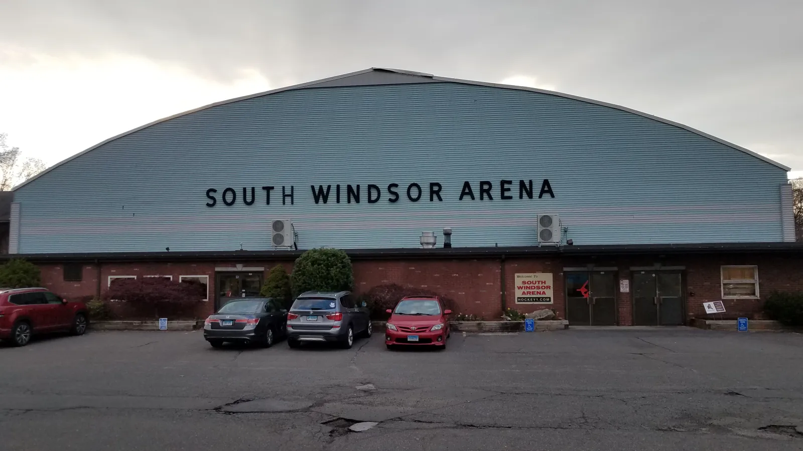 Front facade of South Windsor Arena: a barrel-roofed metal building with "SOUTH WINDSOR ARENA" lettering above the brick entrance, parking lot in front, no people. — South Windsor Arena