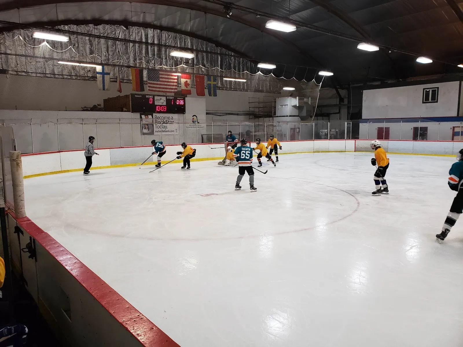 A game in play on a wide sheet seen over a red-topped board, players in yellow and white spread across the ice under a dark arched roof. — South Windsor Arena