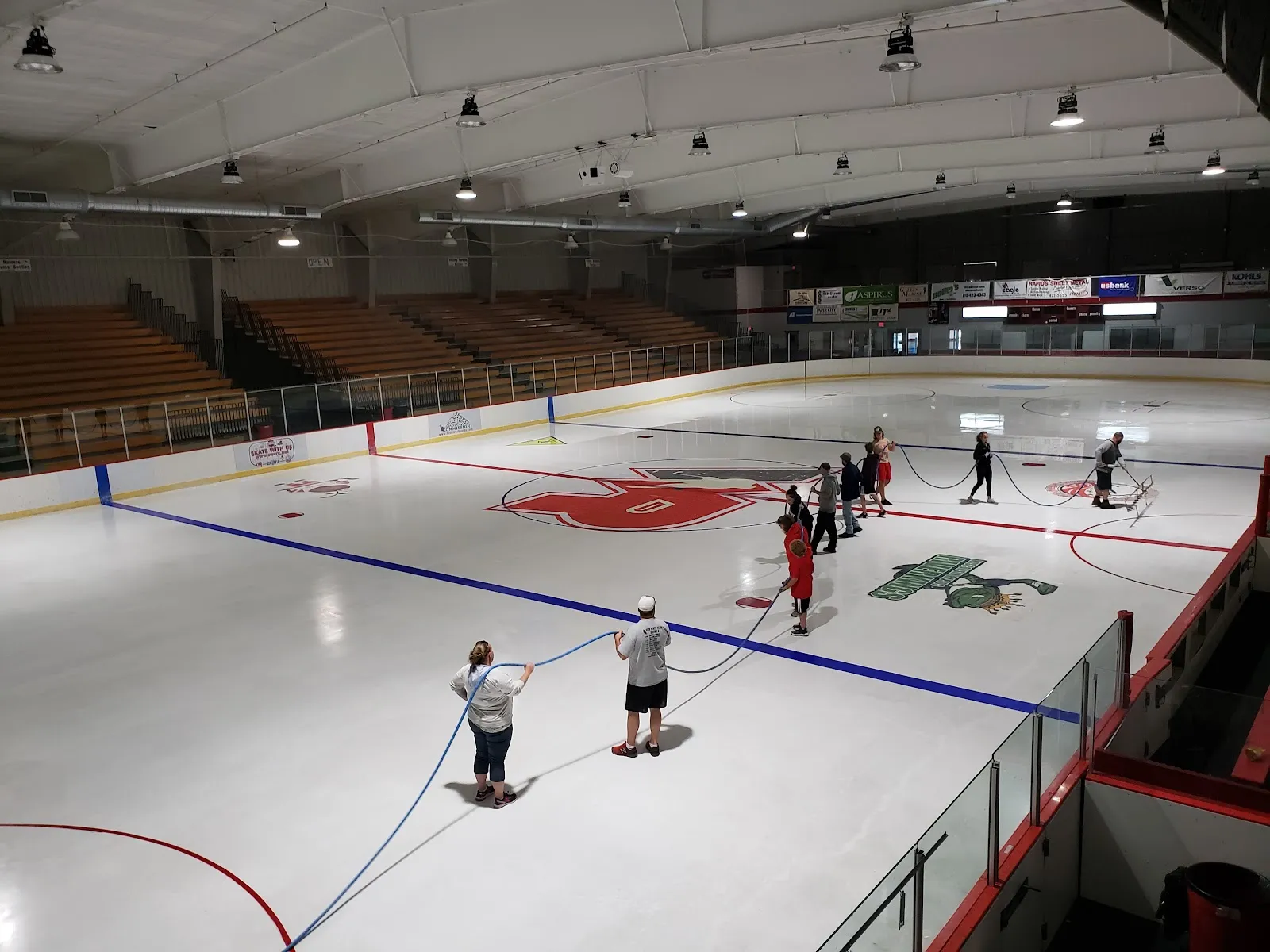 The ice sheet at South Wood County Recreation Center with the red and blue lines, the centre logo and the grandstand beyond — South Wood County Recreation Center