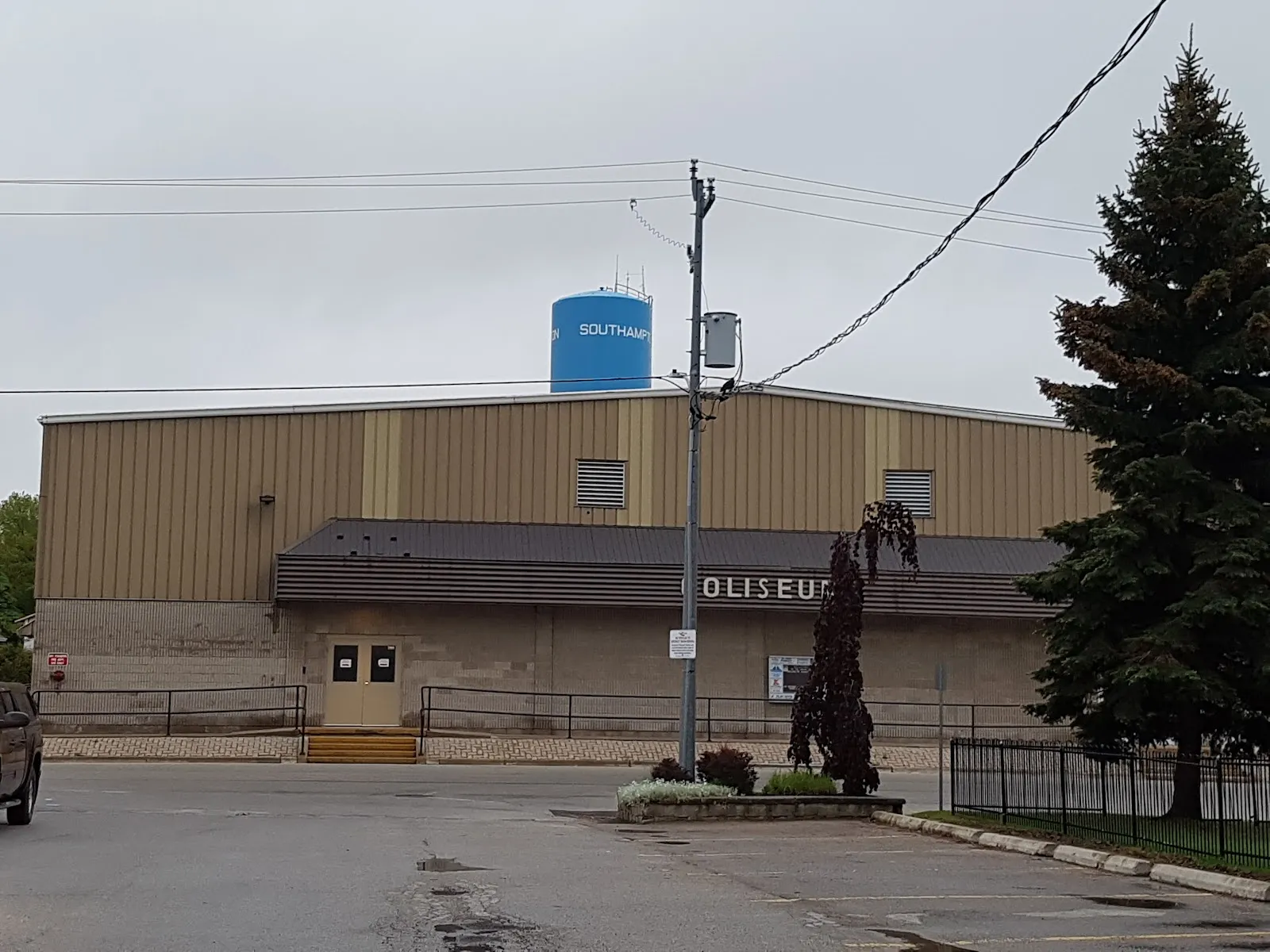 The tan metal-clad side of Southampton Coliseum with a dark canopy over its door and the lettering of its name, beside a tall evergreen and under the town's blue water tower, seen across the parking lot. — Southampton Coliseum