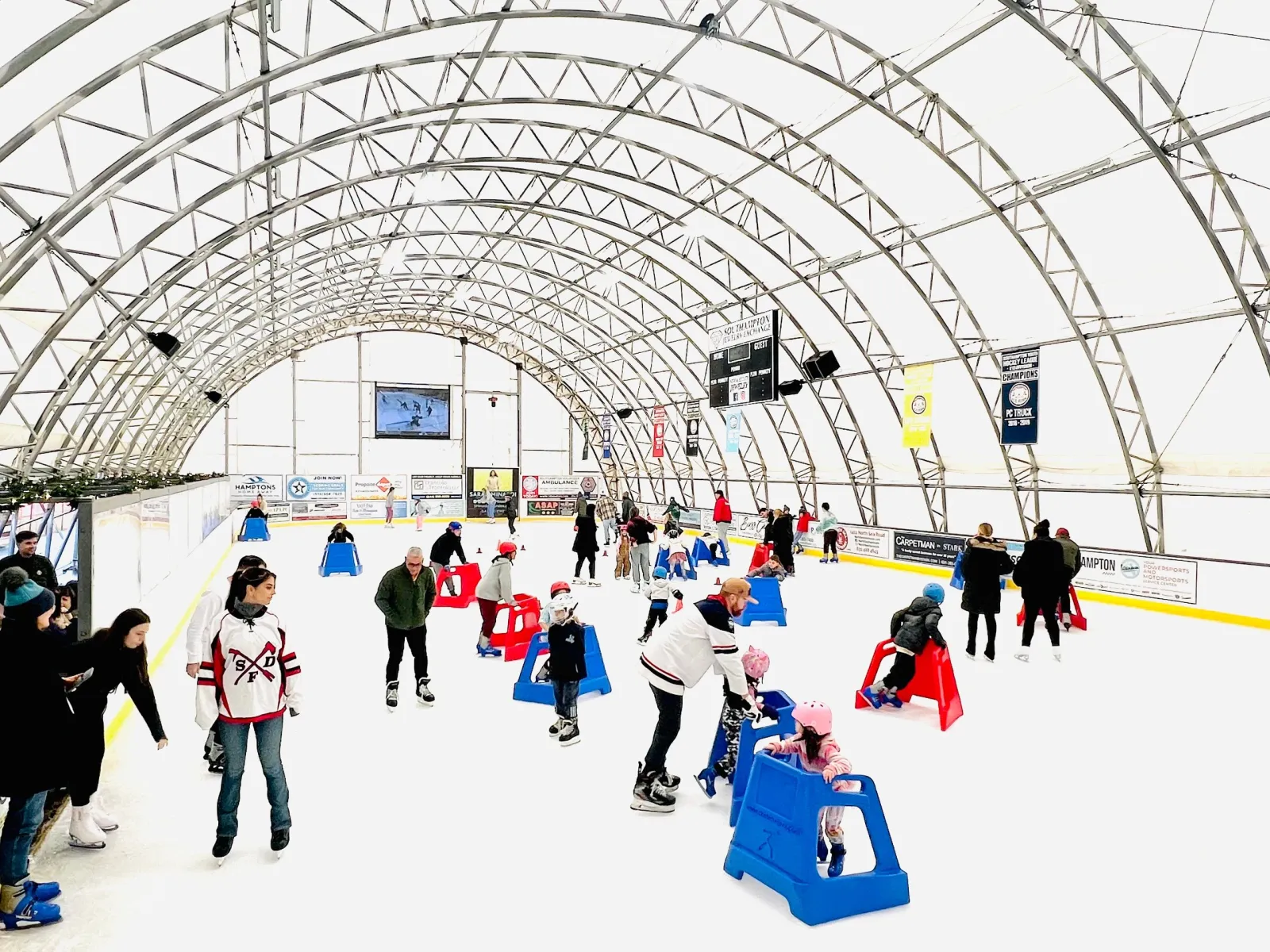Una sesión de patinaje público bajo un techo arqueado de tela, con patinadores repartidos por el hielo entre conos y una bandera y banderines de la pista en la pared del fondo. — Southampton Ice Rink