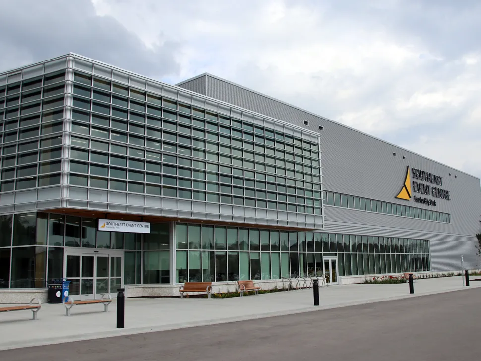La fachada de vidrio y paneles metálicos del Southeast Event Centre sobre Elmdale Street, con el logotipo dorado del centro en la pared, bancas a lo largo del andador de entrada y el largo volumen de la arena detrás. — Southeast Event Centre