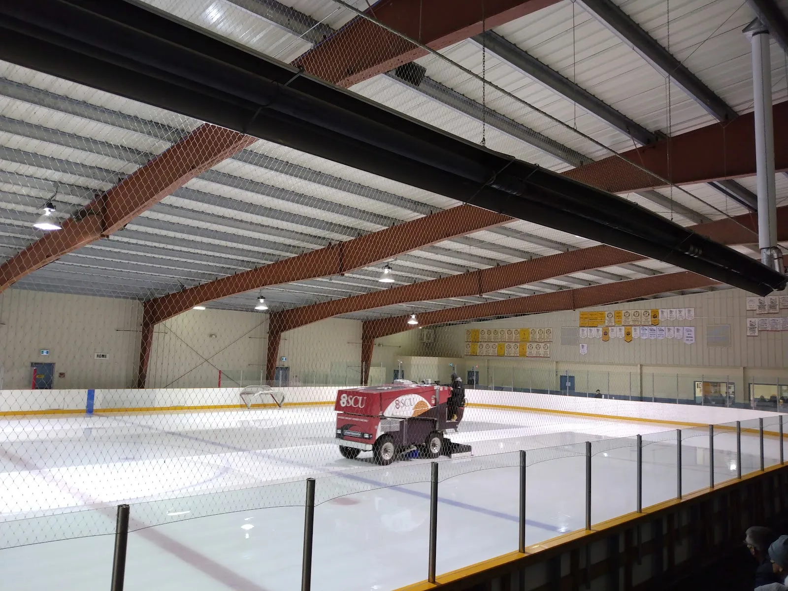 Una pulidora de hielo terminando de renovar la superficie de una de las dos pistas de hielo del centro, vista a través de la red de protección, bajo vigas rojas de acero del techo con mantas de campeonato en la pared del fondo. — Southeast Event Centre