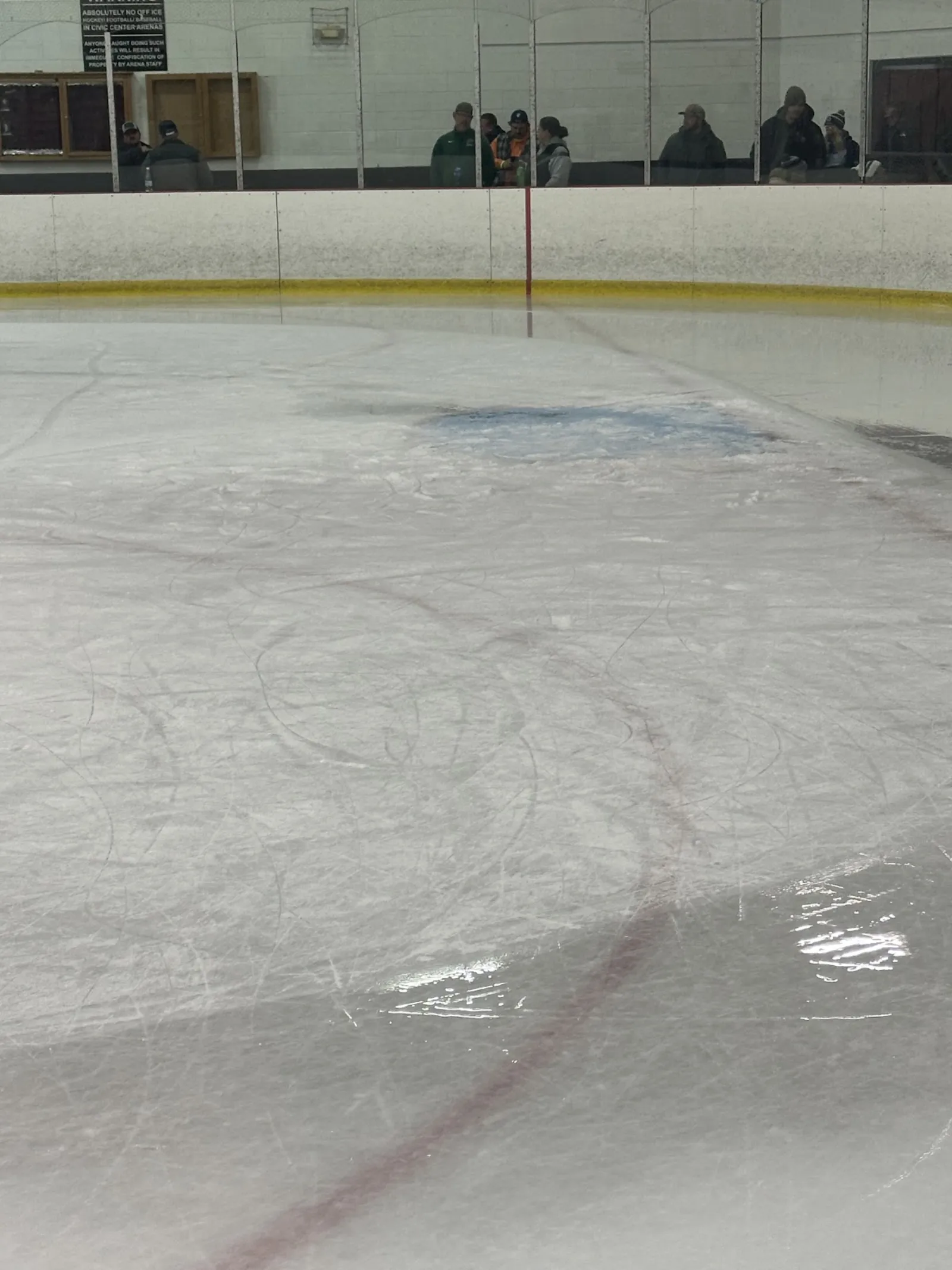 A scarred, scraped ice sheet seen from rink-side, with a sign reading "...IN CIVIC CENTER ARENAS" mounted on the boards and a handful of distant bystanders at the glass in the background. — Southgate Arena