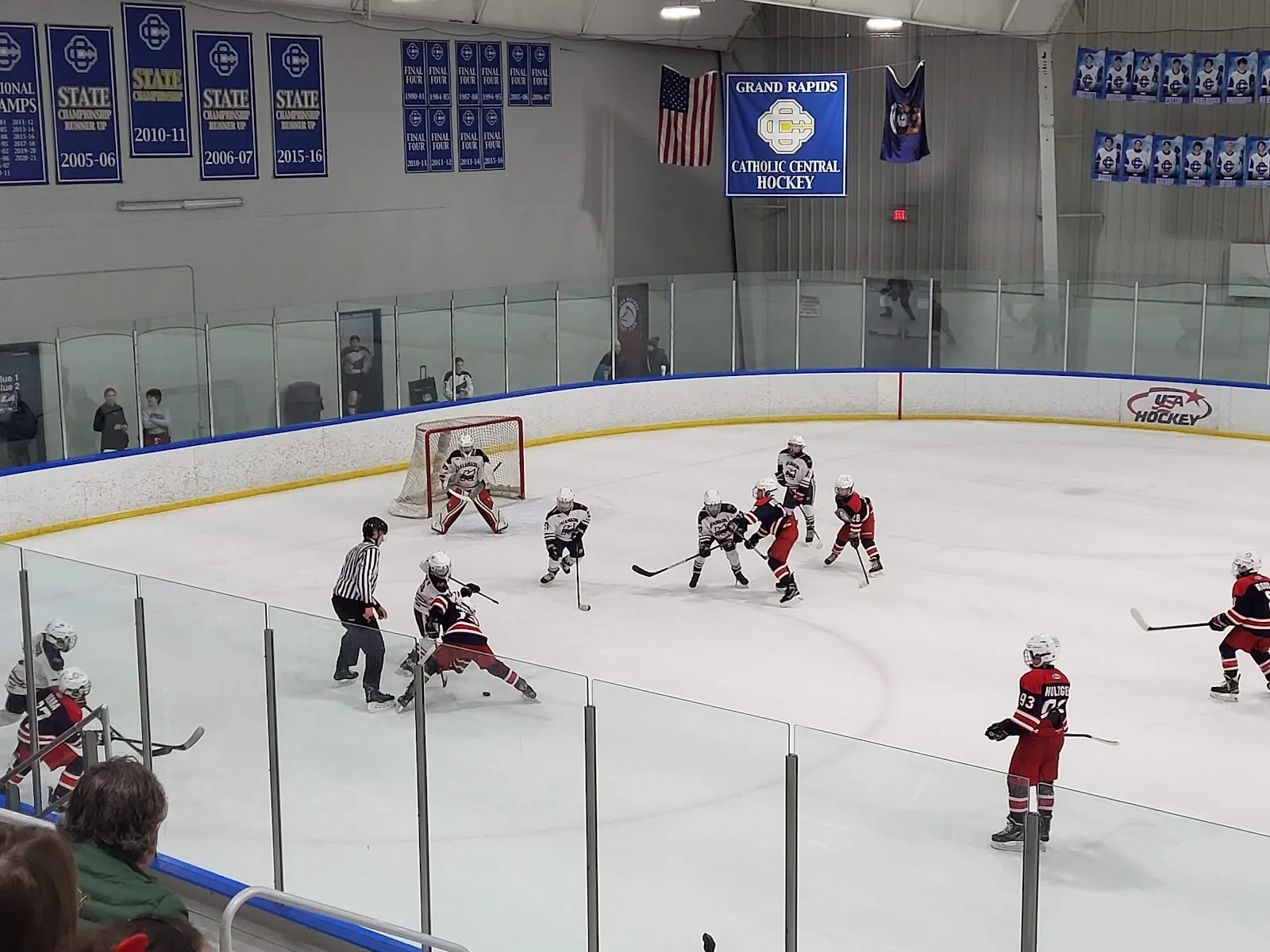 A game seen from the stands over a clear rail, players spread across the ice beneath rows of blue championship banners on a grey wall. — Southside Ice Arena
