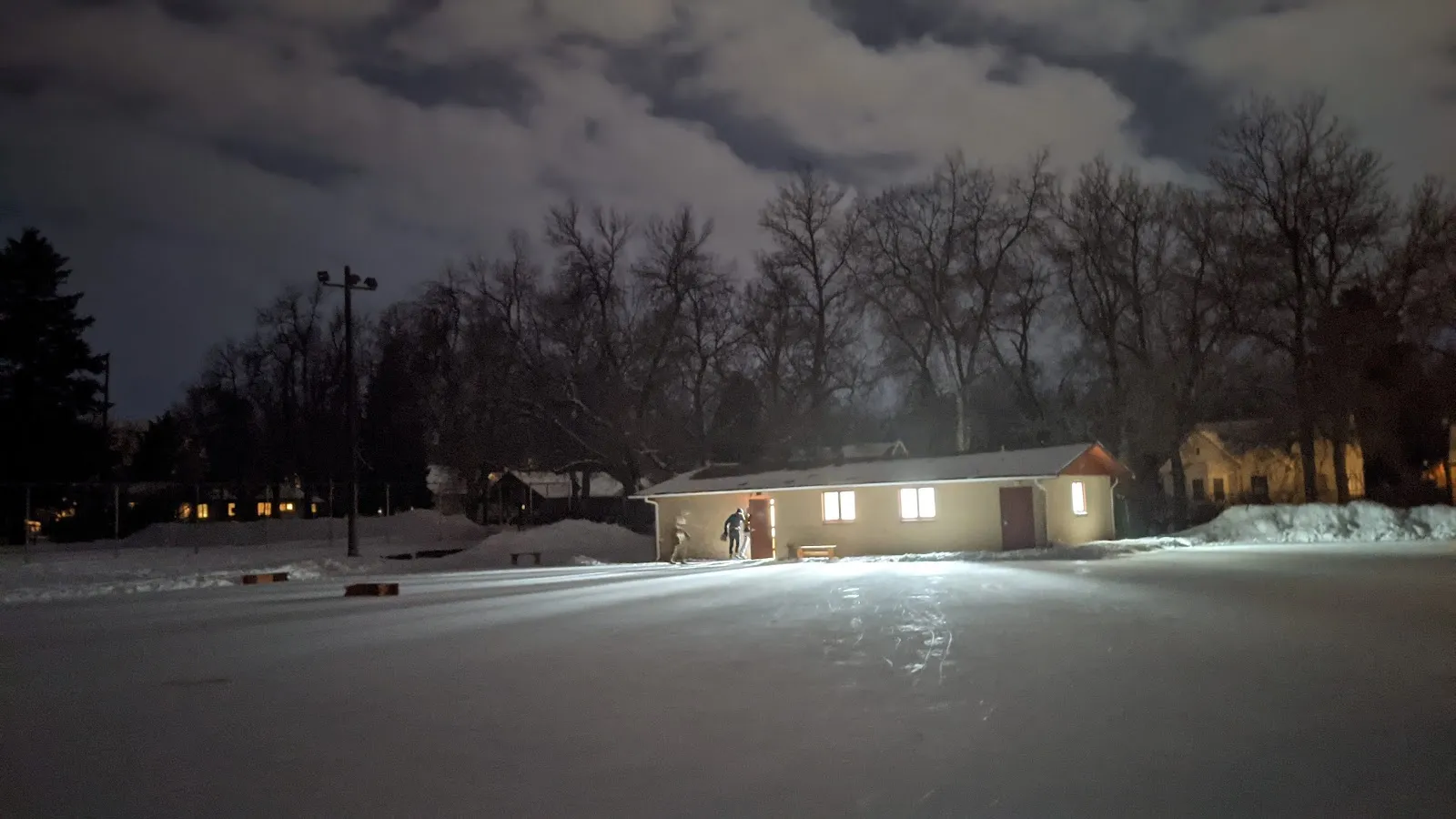 The low warming house at the edge of the lit rink at Southside Park at night, with lit windows, snow banks and bare trees behind. — Southside Park Ice Rinks