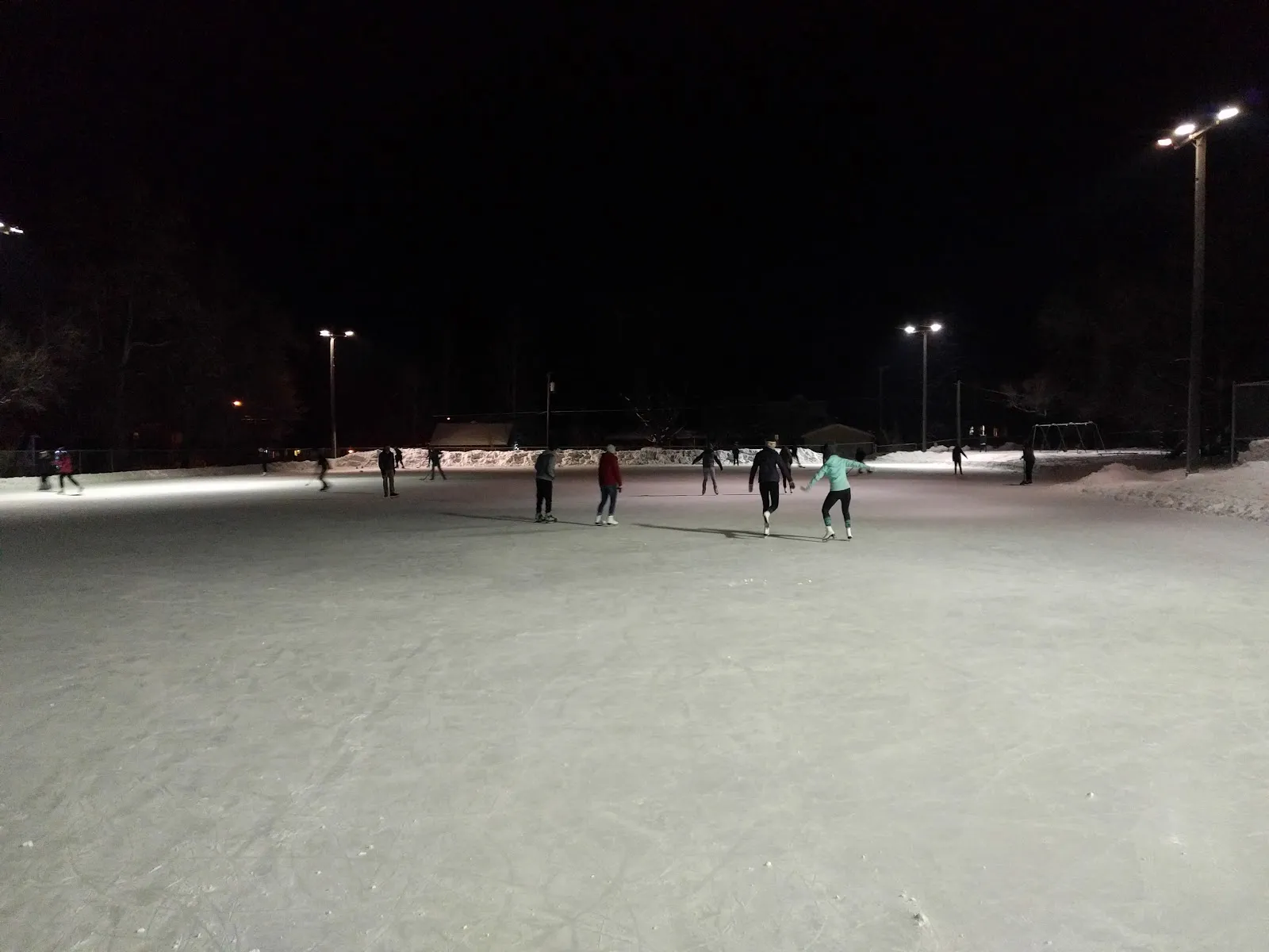 A public skating night on the lit outdoor ice at Southside Park, with skaters at a distance, floodlights on poles and snow banks around the sheet. — Southside Park Ice Rinks