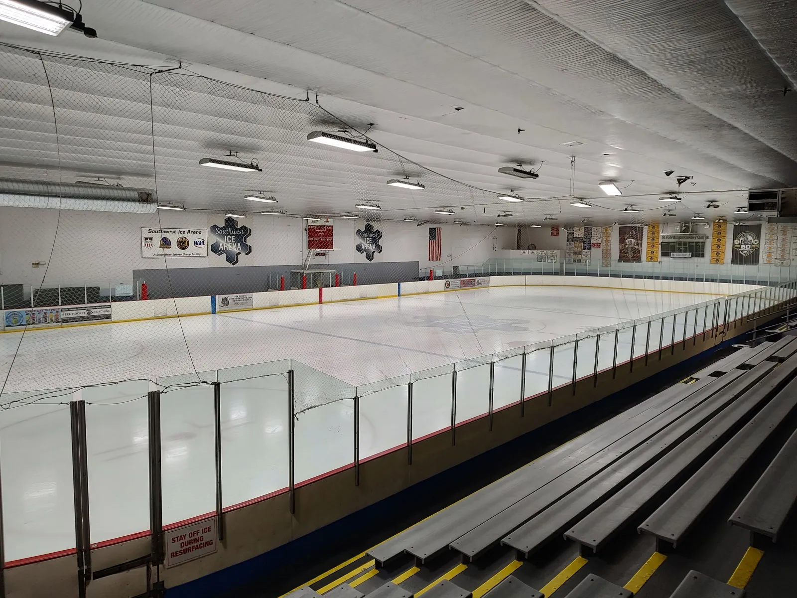 Empty ice sheet viewed from the stands, with the 'Southwest Ice Arena' hexagonal logo on the far wall, team banners along the boards, and protective glass and netting around the rink. — Southwest Ice Arena