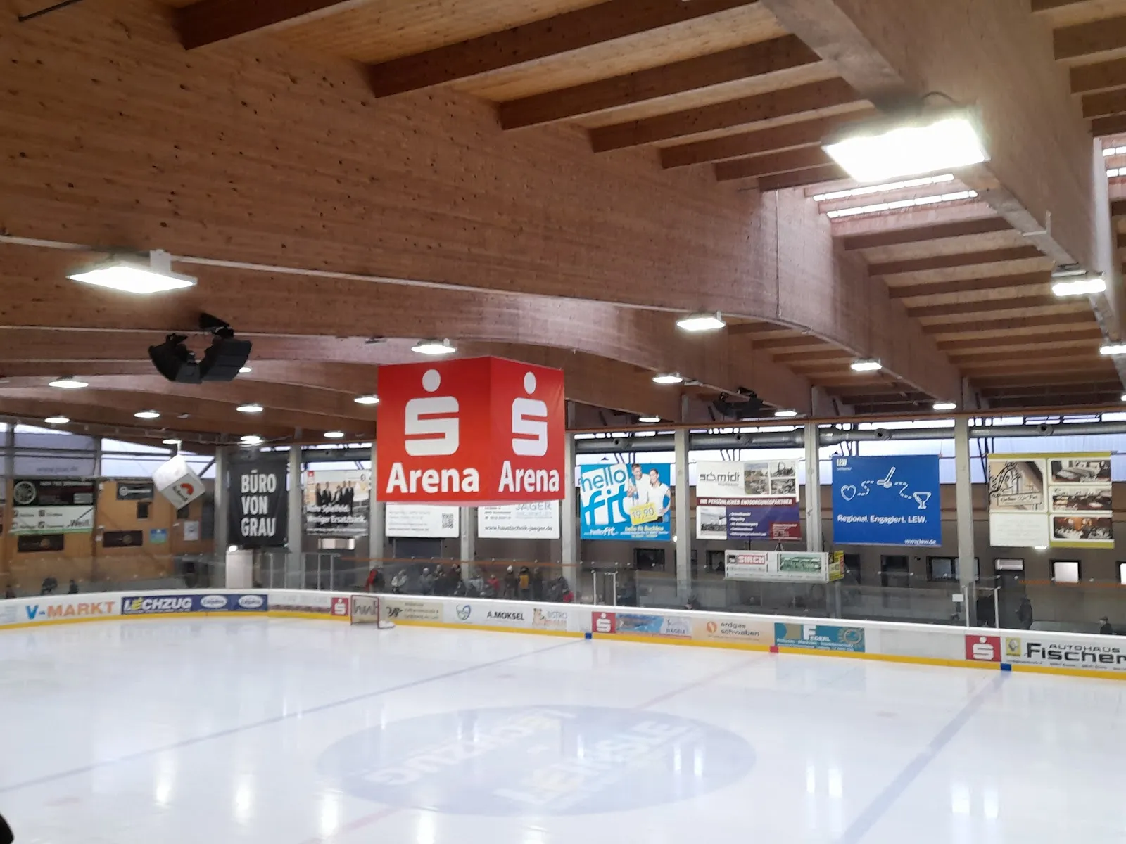The empty ice sheet with a centre-ice logo, sponsor boards along the far side and a red Sparkassen Arena banner hanging from the timber-beamed roof. — Sparkassenarena Buchloe