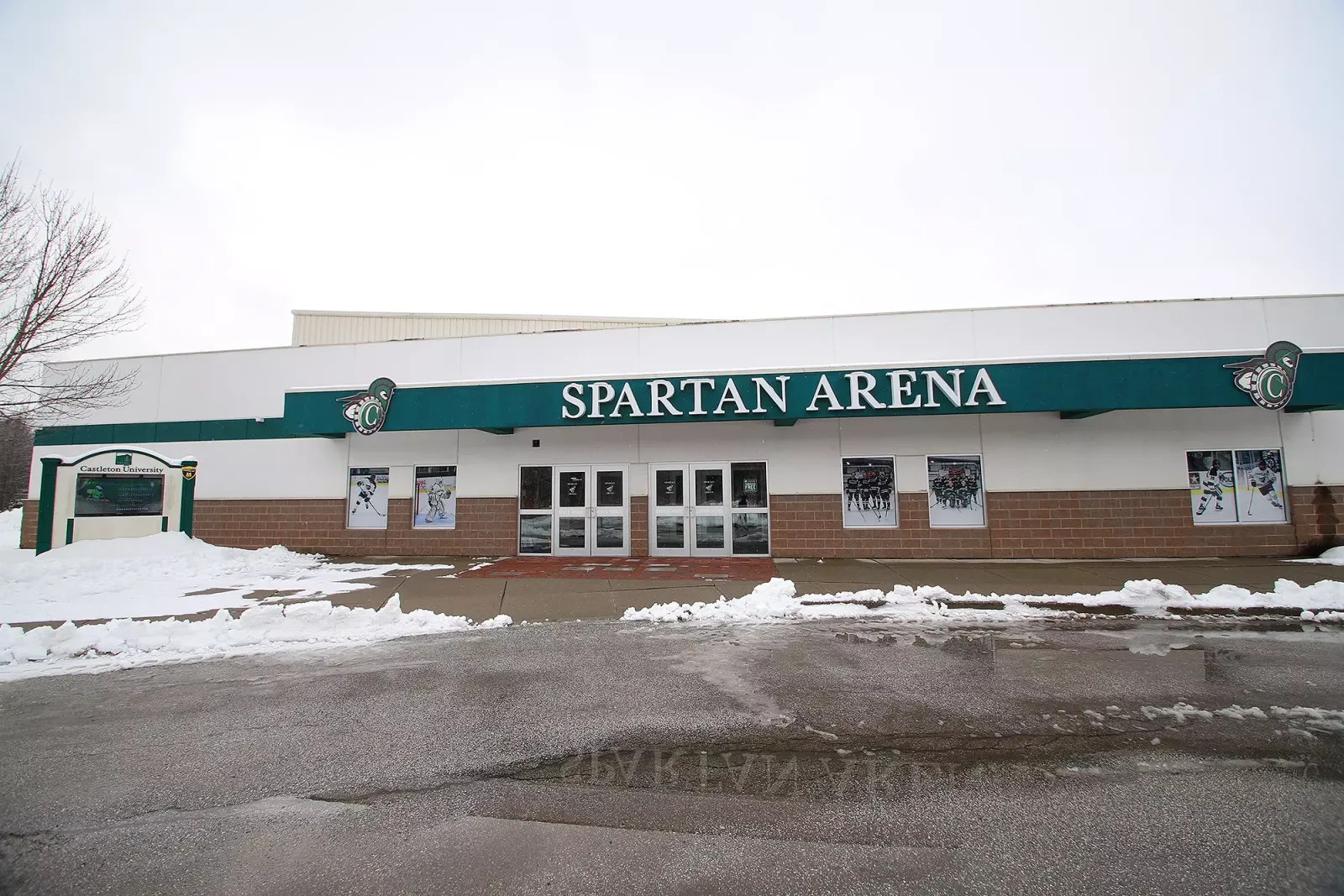 Fachada frontal de Spartan Arena en invierno, con nieve retirada a lo largo del andador, una franja verde con el letrero de 'SPARTAN ARENA' sobre las puertas de la entrada, un letrero monumental de 'Castleton University' en la esquina izquierda y gráficos de acción de hockey en las ventanas a lo largo de la base de ladrillo. — Spartan Arena