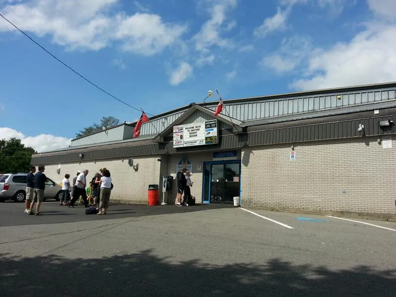 The arena's brick-and-steel front with a pitched entrance canopy, notice sign and flags, and a few people gathered by the door on a sunny day. — Spencerville Arena