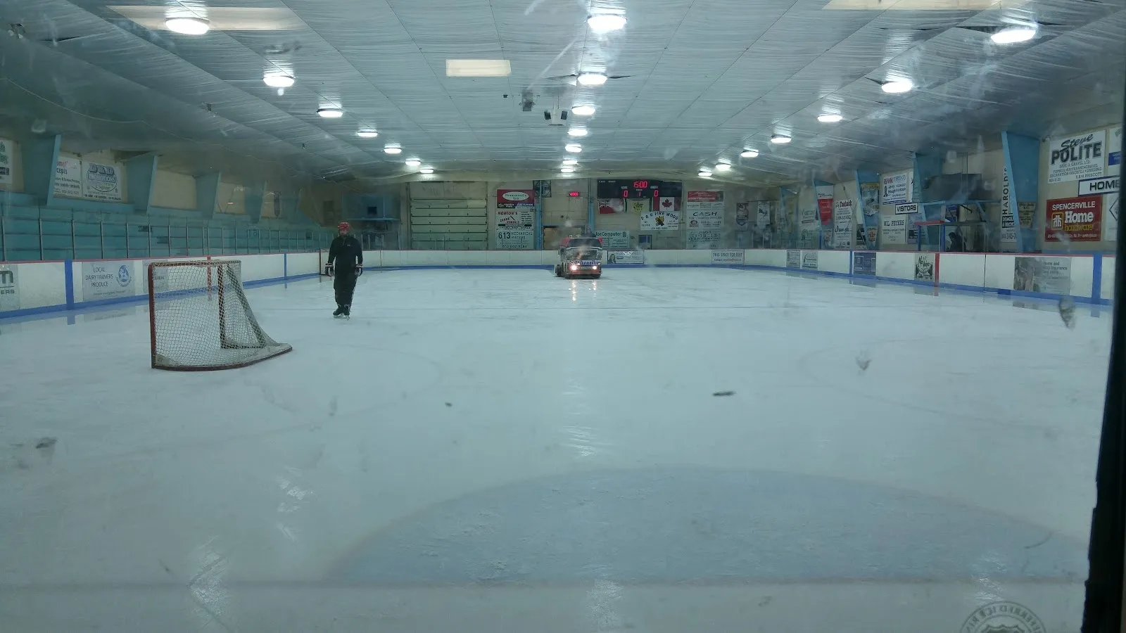 A wide empty sheet with a net at the left and a long blue-green wall behind, a few people far along the boards under a bright ceiling. — Spencerville Arena