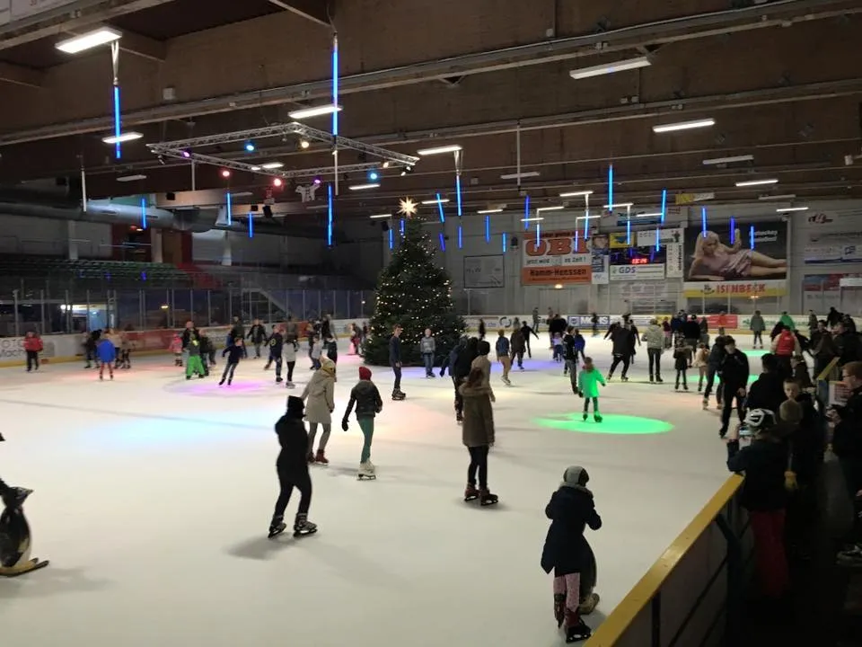 Eine gut besuchte öffentliche Laufzeit in der Eissportarena Hamm, Schlittschuhläufer über dem Eis mit farbigen Bodenlichtern und einem Weihnachtsbaum an der Bande. – Spengel Eissportarena