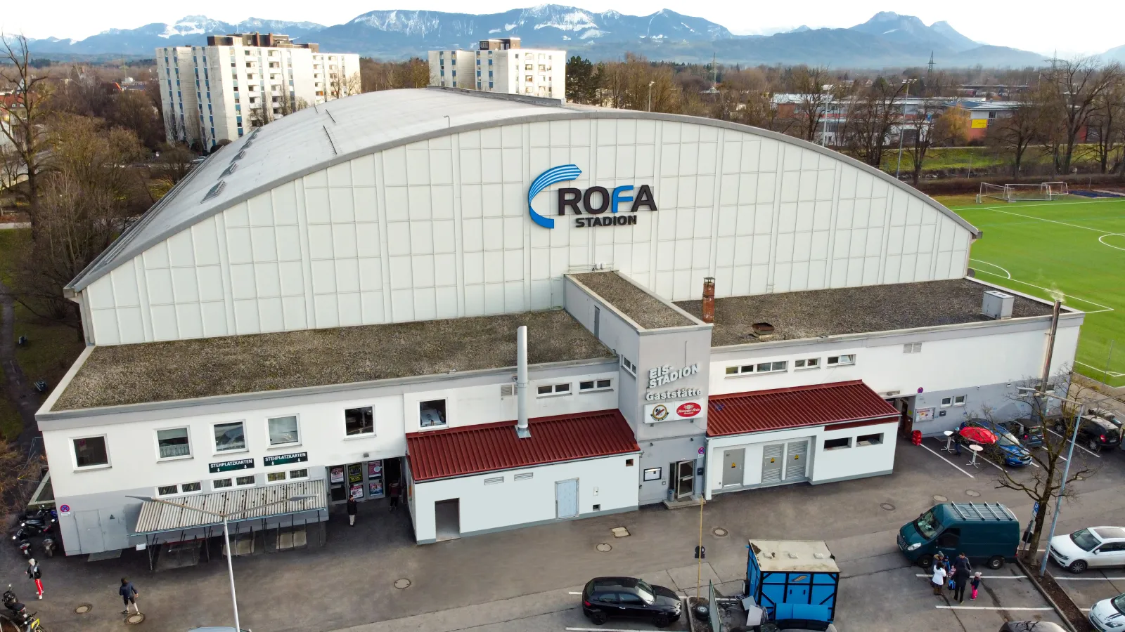 The white barrel-roofed ice stadium seen from above, with its entrance block, the car park in front and the Alps behind. — SPIE Arena Rosenheim