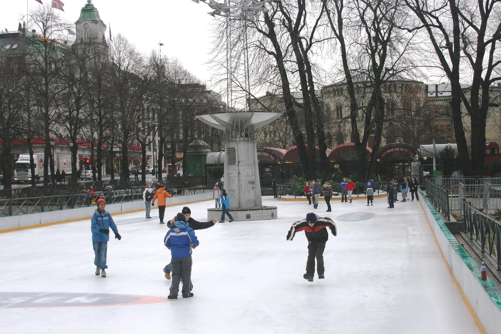 Skøyteløpere på friluftsbanen Spikersuppa i sentrum av Oslo, med bare trær og et monument bak vantene. — Spikersuppa