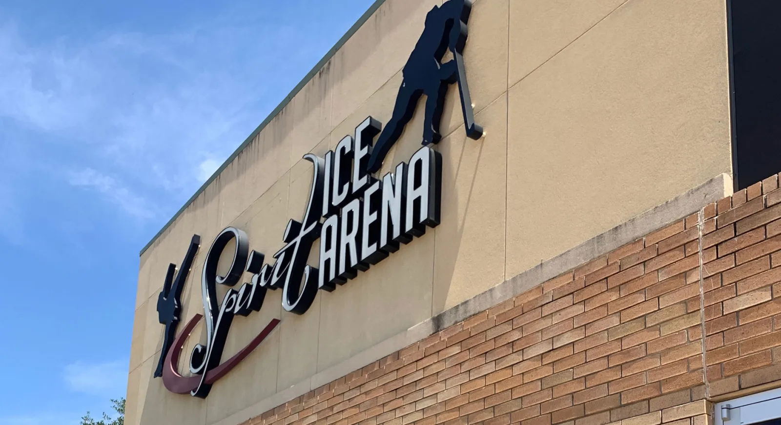 Acercamiento al letrero iluminado de "Spirit Ice Arena" y a la silueta de un jugador de hockey en la fachada color arena del edificio, contra un cielo azul — Spirit Ice Arena
