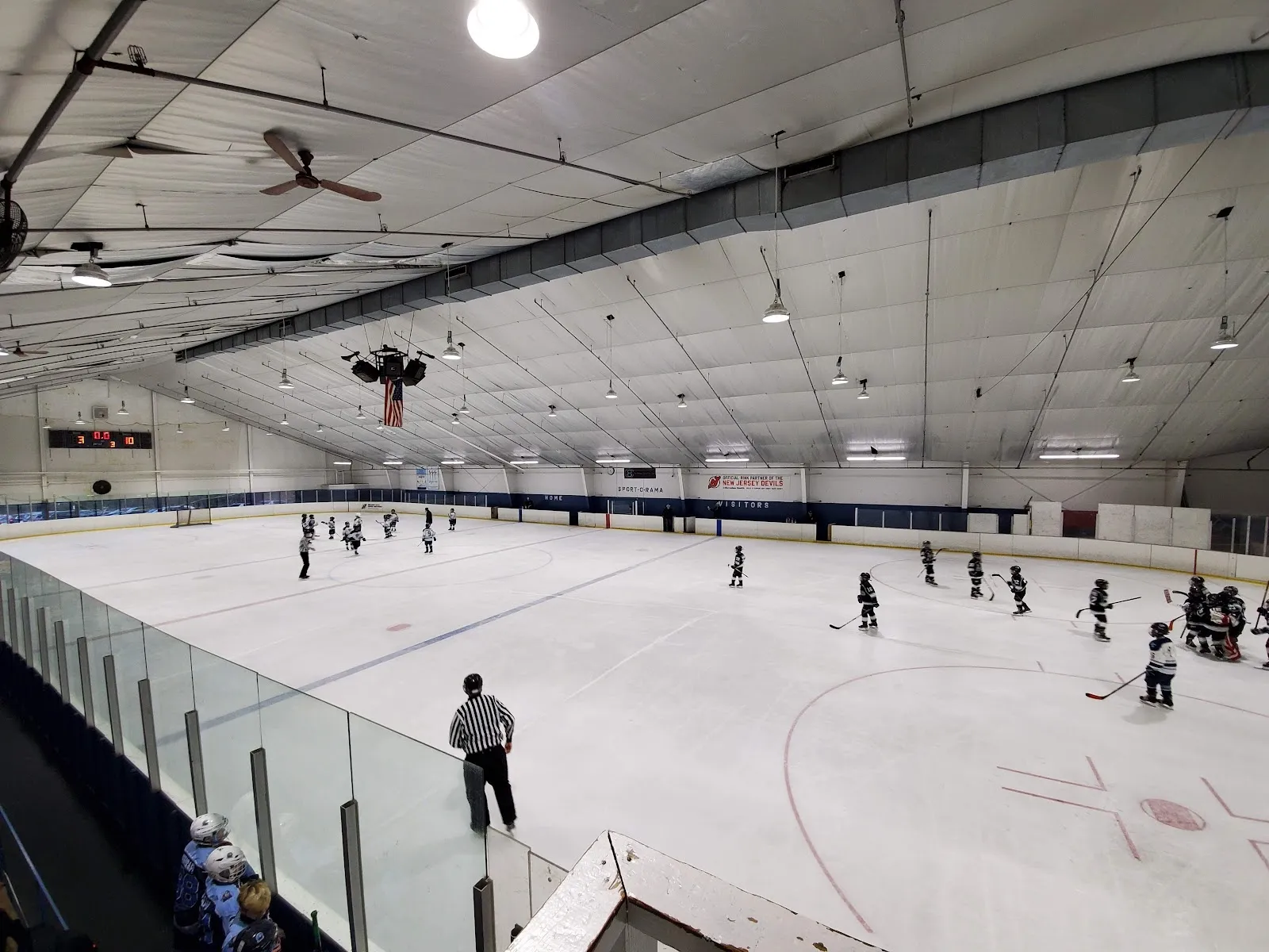 A wide view down a glass-topped rail of a sheet with players spread across the ice under a pale roof with a ceiling fan. — Sport-O-Rama Ice Rinks