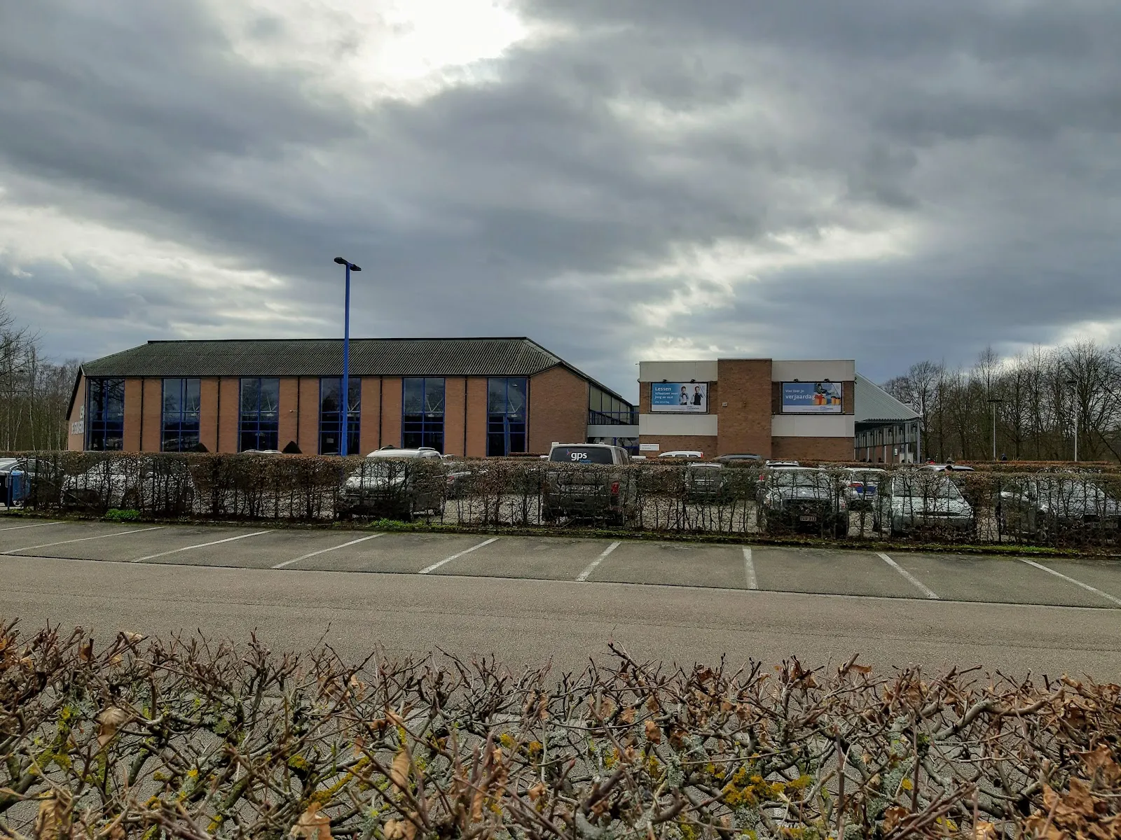 Le centre sportif vu du stationnement : une longue halle de brique au toit sombre avec une enseigne sur la façade de droite, et la haie devant. — Sport Vlaanderen Hasselt