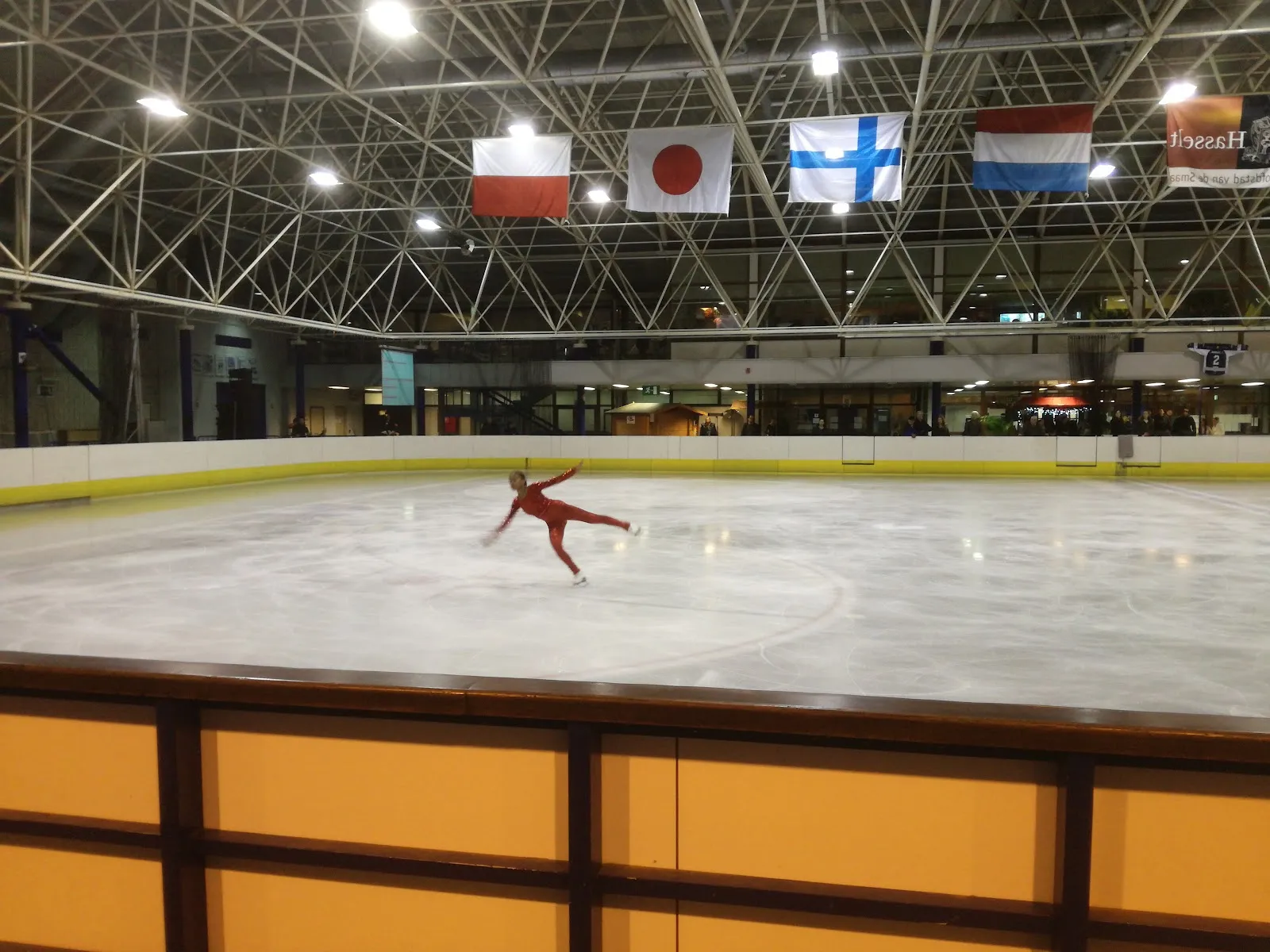 La patinoire couverte sous le toit d’acier avec les drapeaux nationaux suspendus au-dessus et les bandes jaunes ; un patineur artistique, loin sur la glace. — Sport Vlaanderen Hasselt