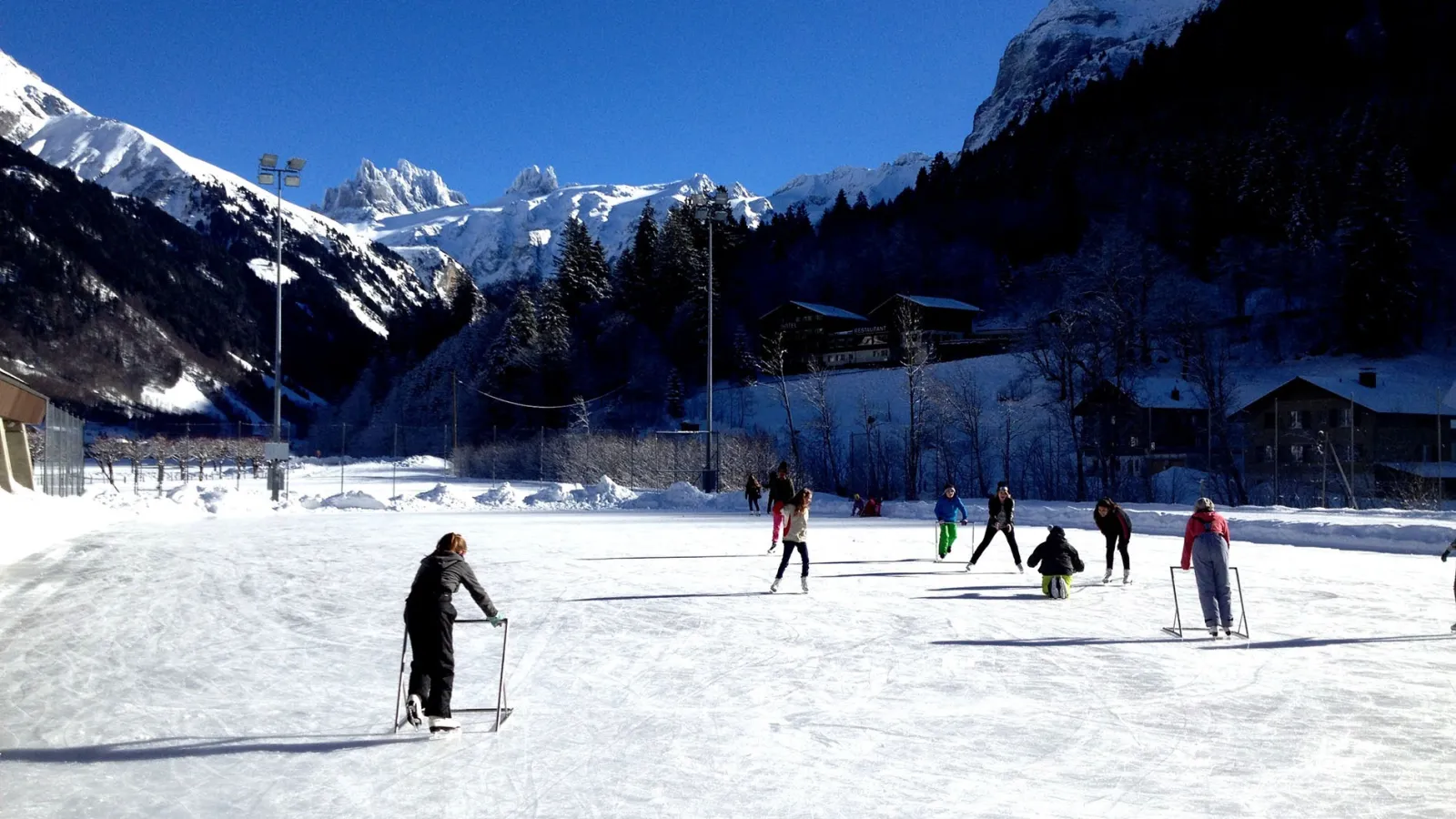 Skaters on the natural-ice rink at Engelberg, snow-covered slopes and the mountain wall rising behind the boards. — Sporting Park Engelberg
