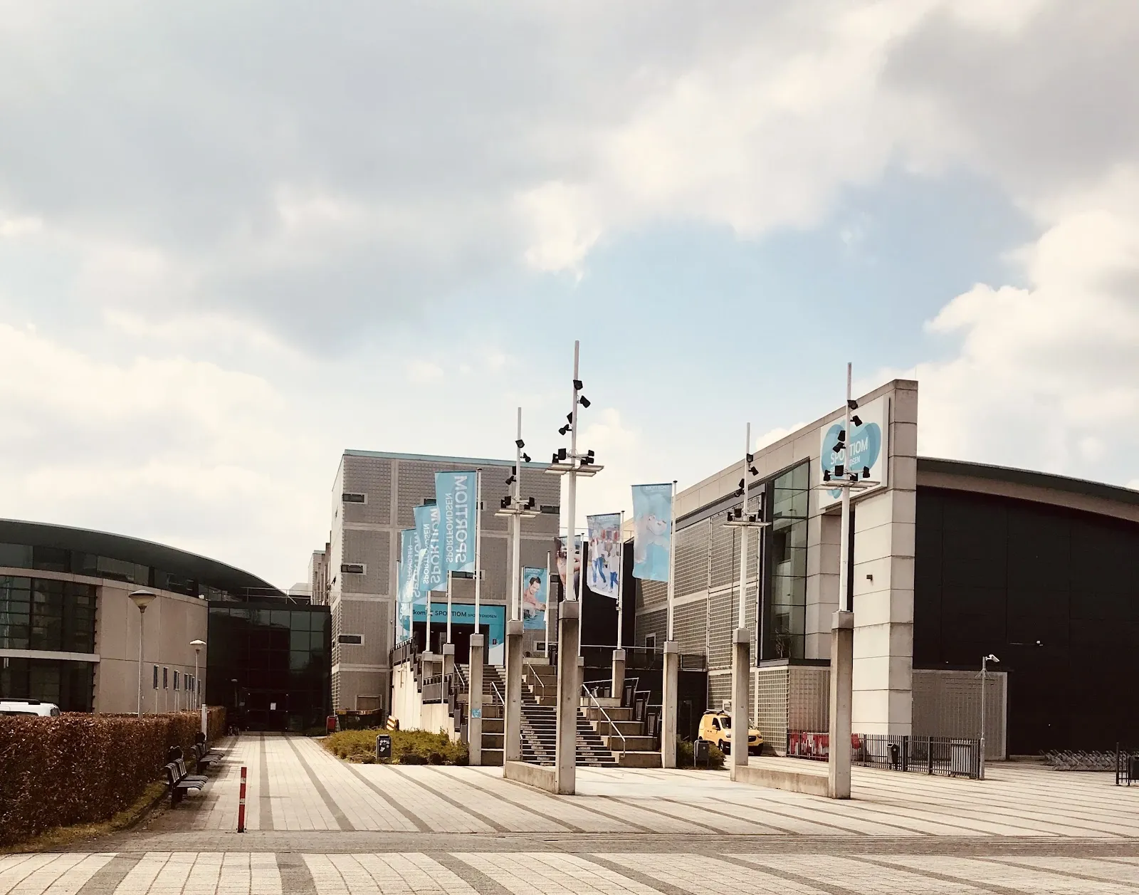 The paved approach to Sportiom in 's-Hertogenbosch with its tall banner flags, the stepped entrance and the grey-clad hall beside it — Sportiom