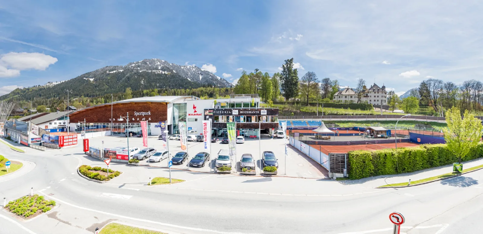 Der Sportpark Kitzbühel von der Straße aus: die lange, geschwungene Holzfront der Sporthalle, der Parkplatz mit Fahnen und die Tennisplätze daneben, dahinter die Berge. – Sportpark Kitzbühel Eishalle