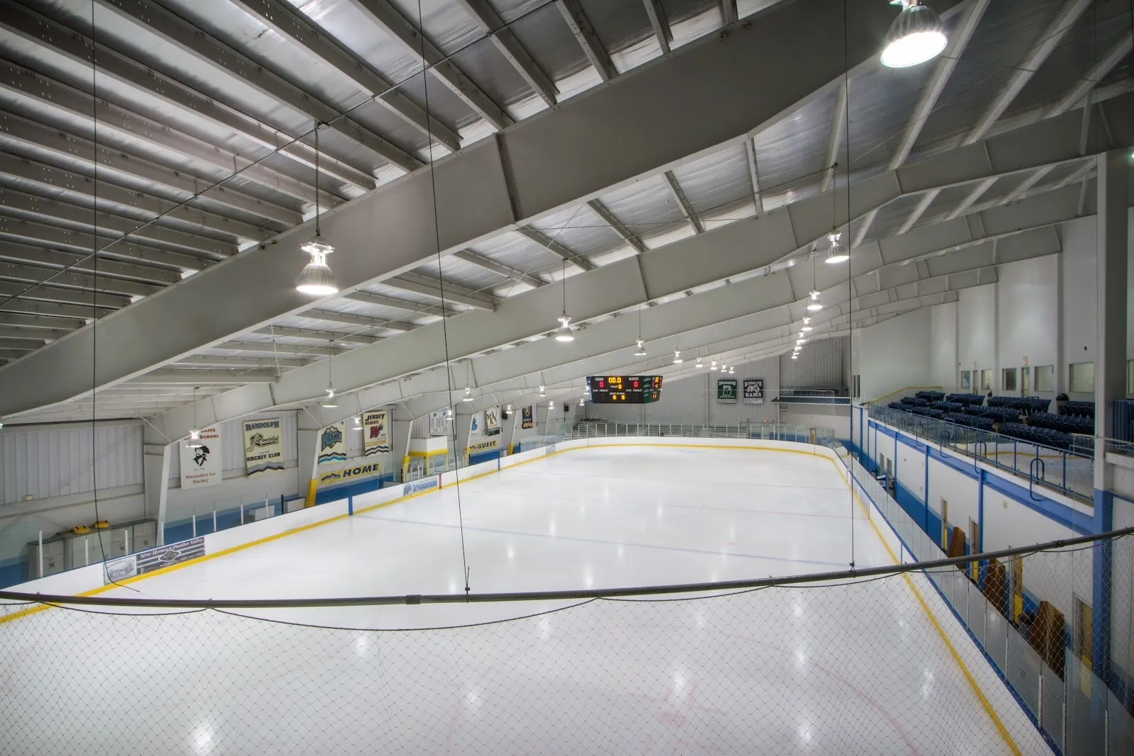 An empty sheet seen from the end boards, white ice with blue edge lines under a long steel-framed roof with blue rail panels. — Sportscare Arena