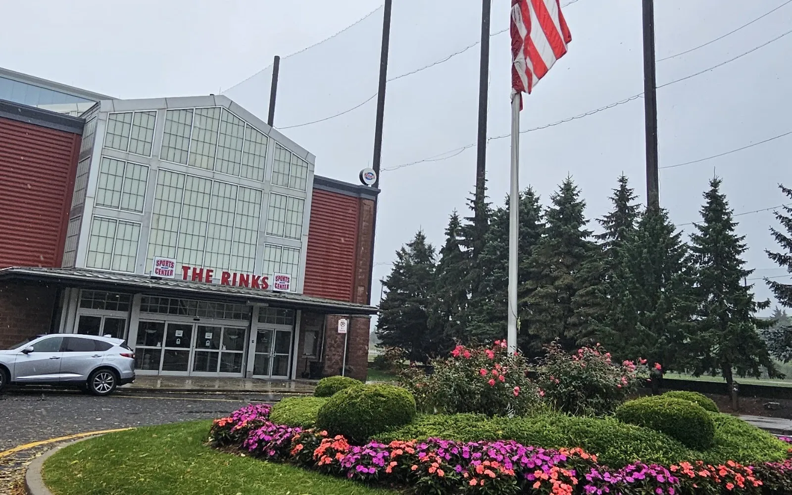 The Rinks entrance at SportsCenter of Connecticut, a glass gable between red-clad walls, with a flower bed and flagpole in front — SportsCenter of Connecticut