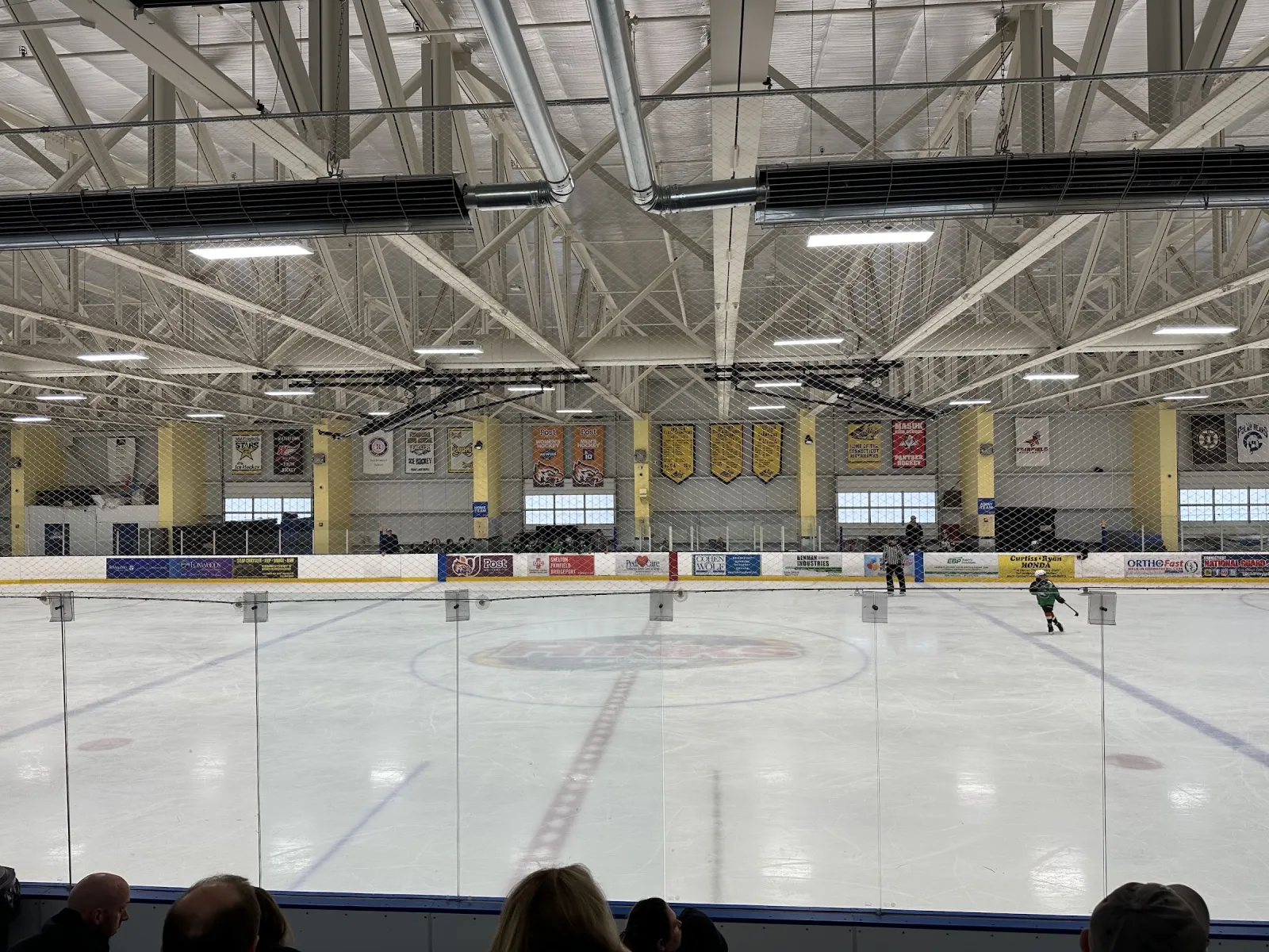 The ice seen from behind the glass, with a lone skater, yellow-and-white banners and a steel-truss roof. — SportsCenter of Connecticut