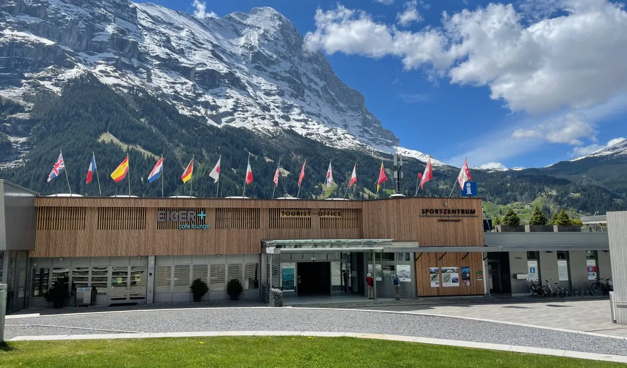 The front of the Sportzentrum Grindelwald with a row of flags and the snow-covered peaks behind it — Sportzentrum Grindelwald