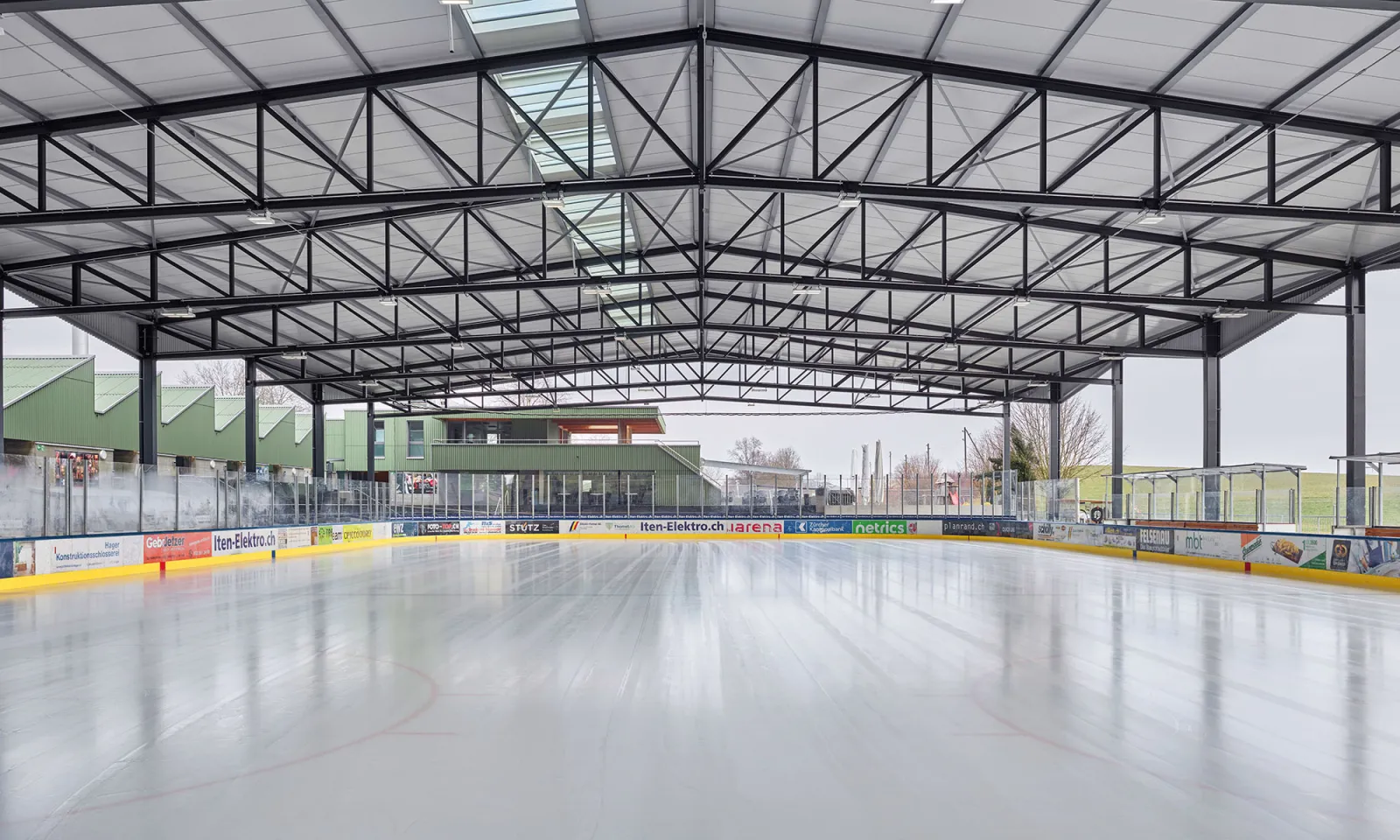 The empty ice under the steel-trussed roof of the covered rink, with the restaurant building behind the boards — Sportzentrum Hirzi