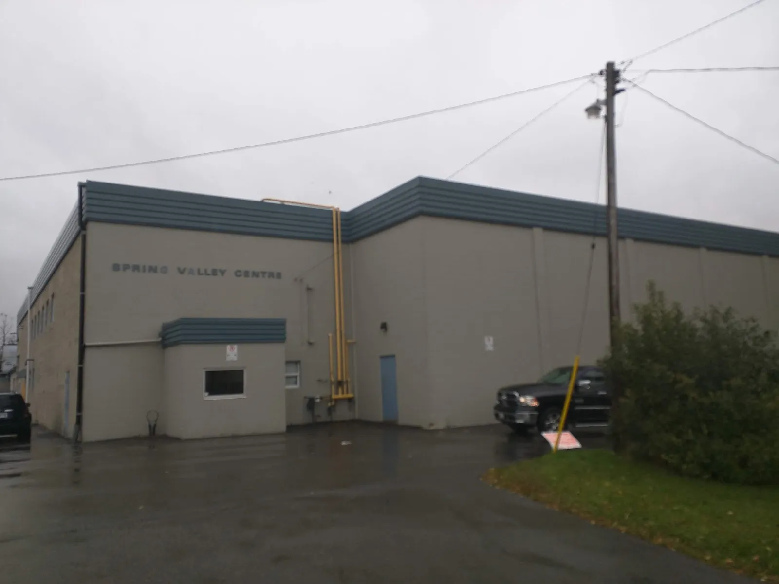 Edificio de la arena de metal color beige con la leyenda "Spring Valley Arena" y un pequeño bloque de entrada, visto desde el estacionamiento en un día gris. — Spring Valley Arena