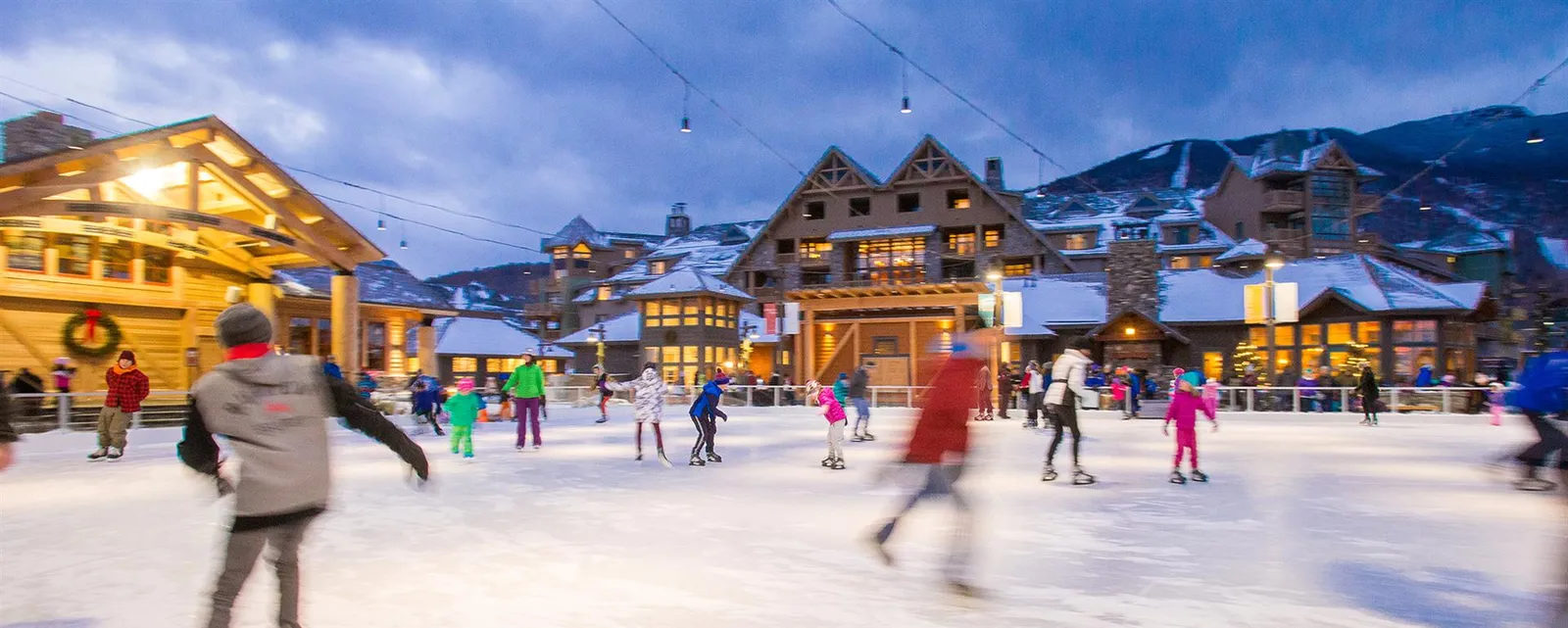 Skaters on the Spruce Peak Village ice rink in Stowe, with the timber lodge buildings and snowy slopes behind. — Spruce Peak Village Ice Rink
