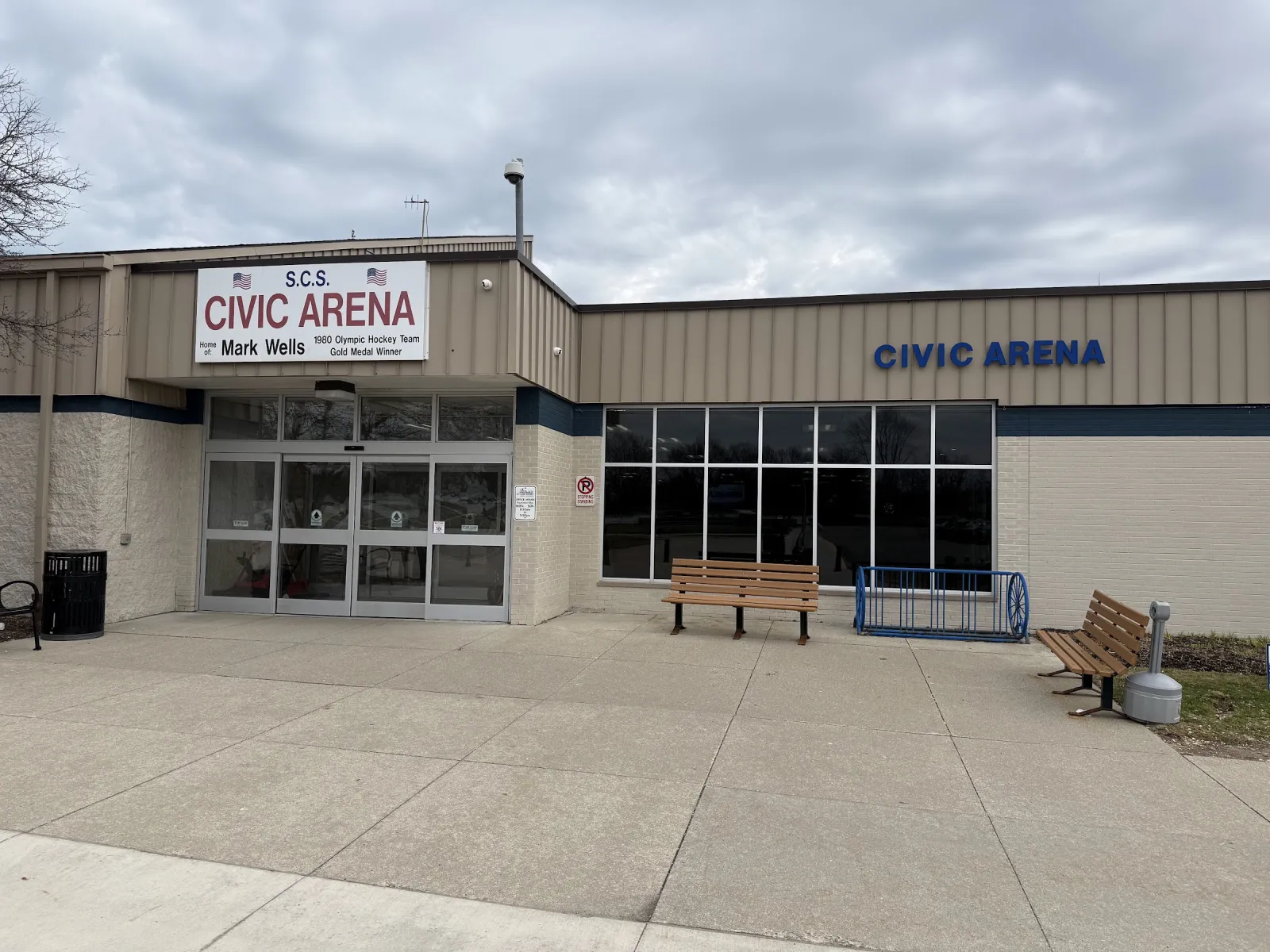 The tan metal building's glass entrance under an S.C.S. Civic Arena sign, with Civic Arena lettered on the wall beside a row of windows and benches out front. — St. Clair Shores Civic Arena