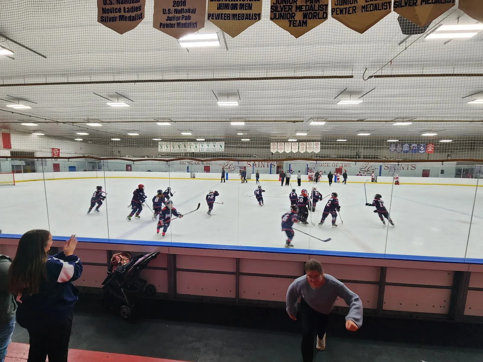 Skaters spread across a wide sheet beneath rows of championship banners, a blue and pink board along the near side. — St. Clair Shores Civic Arena