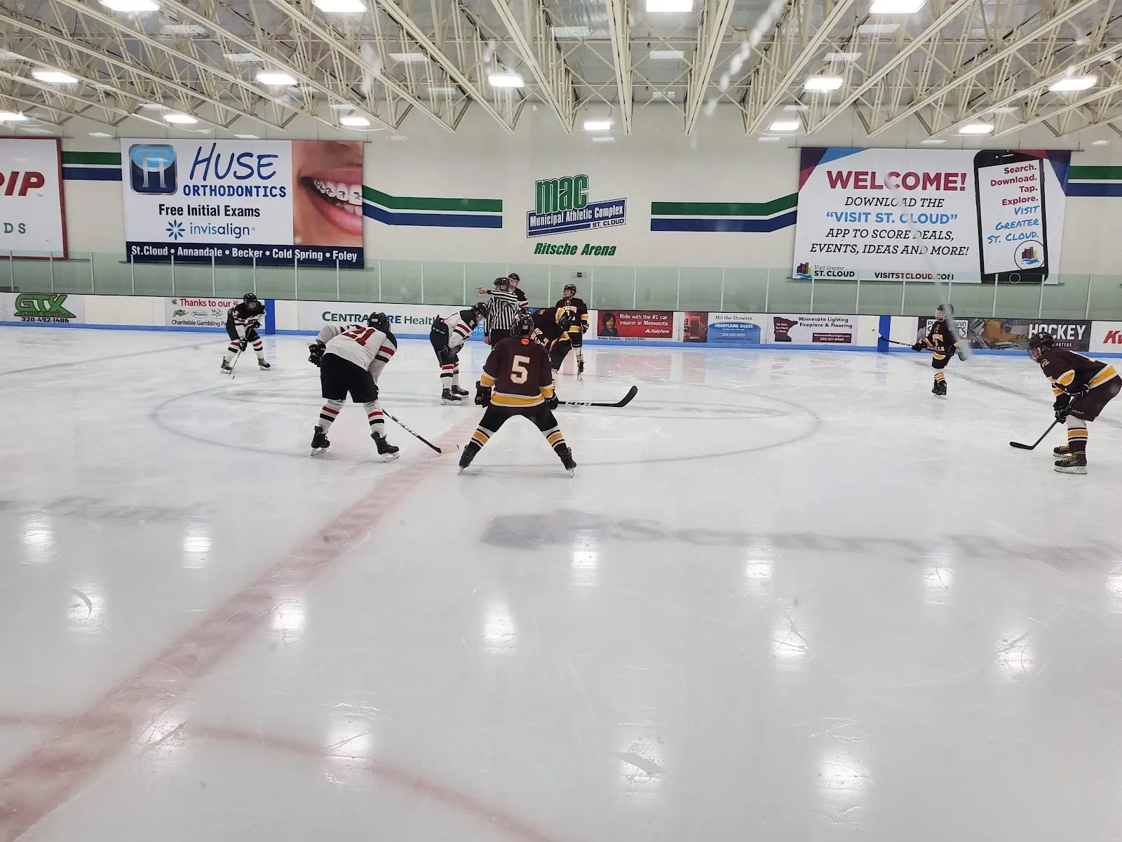 A game under a bright white ceiling, players spread around the faceoff circle on a wide sheet with sponsor boards and welcome banners along the far wall. — Municipal Athletic Complex (MAC)