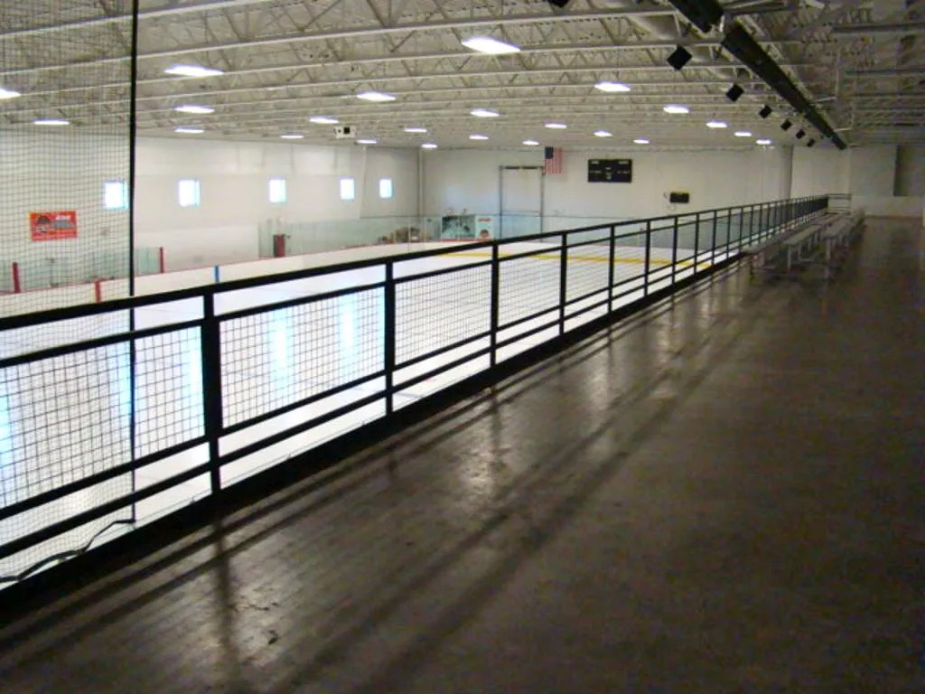 Parte de una pista vista desde el pasillo elevado para espectadores detrás del cristal, con las vallas, las luces del techo y gradas vacías. — St. Croix Valley Recreation Center