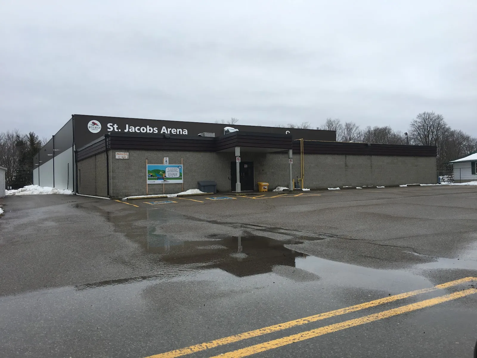 Low dark-clad arena building with a "St. Jacobs Arena" sign above the entrance, seen across a wet parking lot. — St. Jacobs Arena